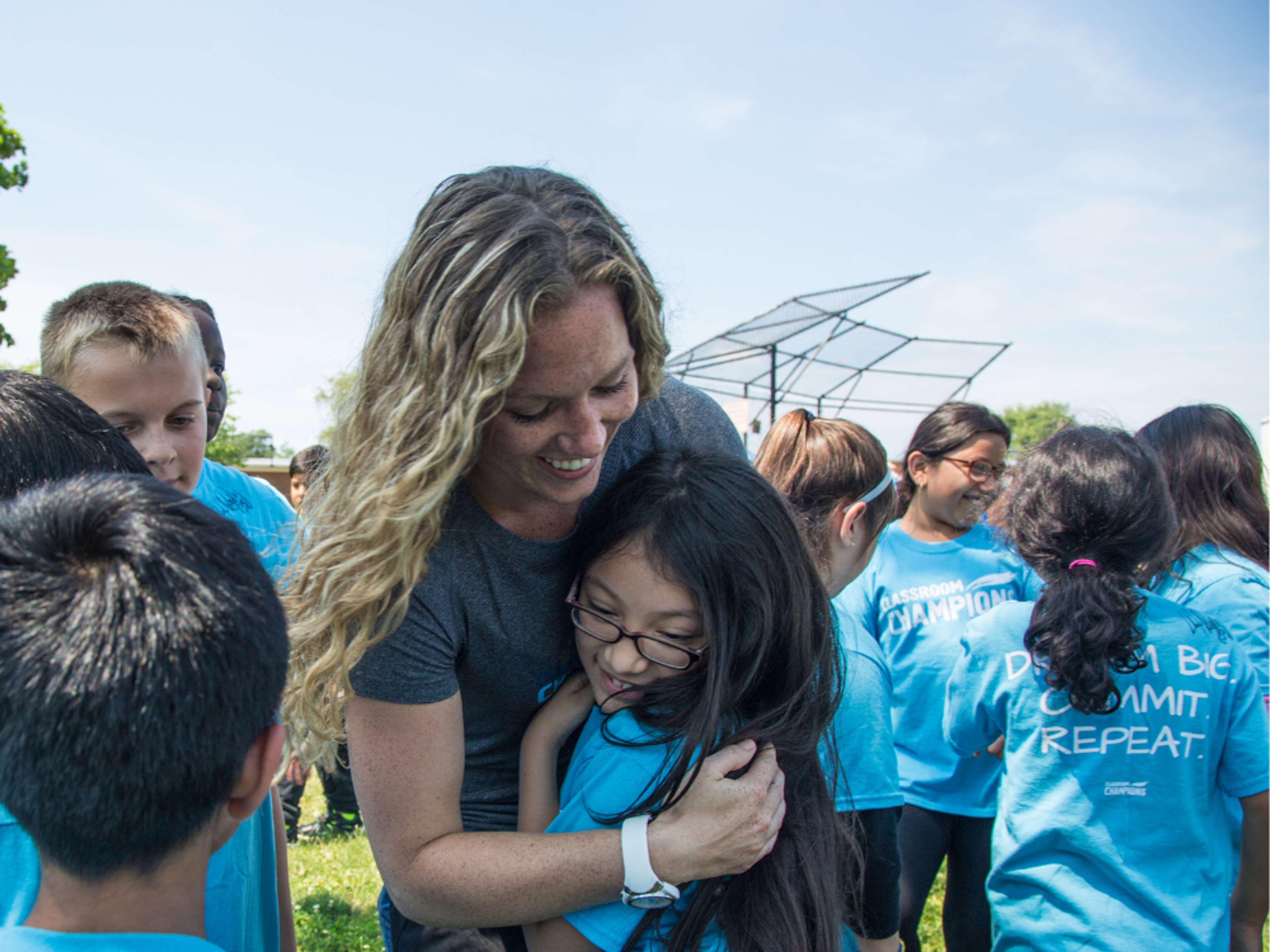 A mentor hugs a student outdoors, surrounded by kids in Classroom Champions T-shirts.