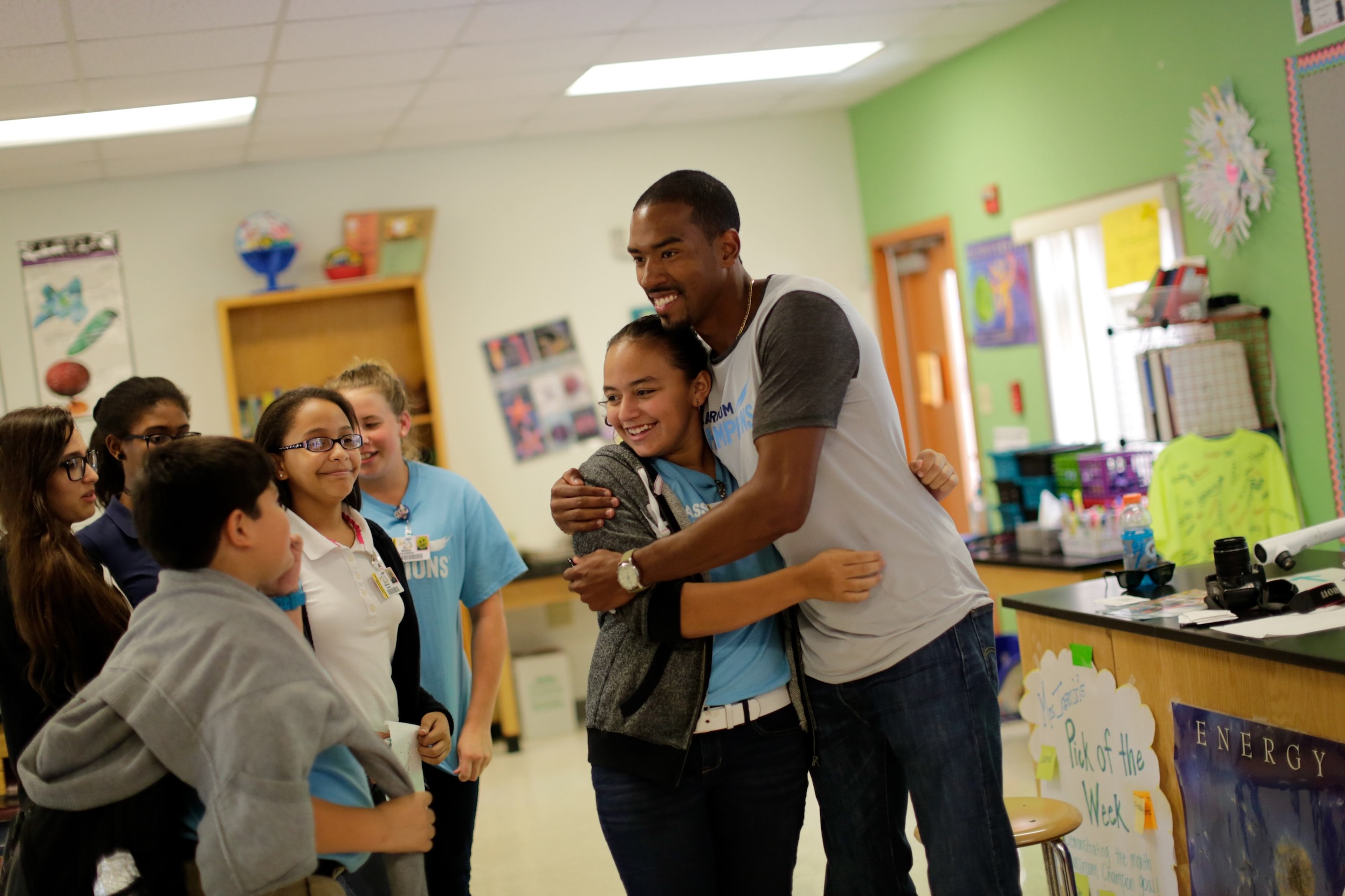 An athlete mentor hugs a student while classmates smile nearby in a Classroom Champions classroom visit.