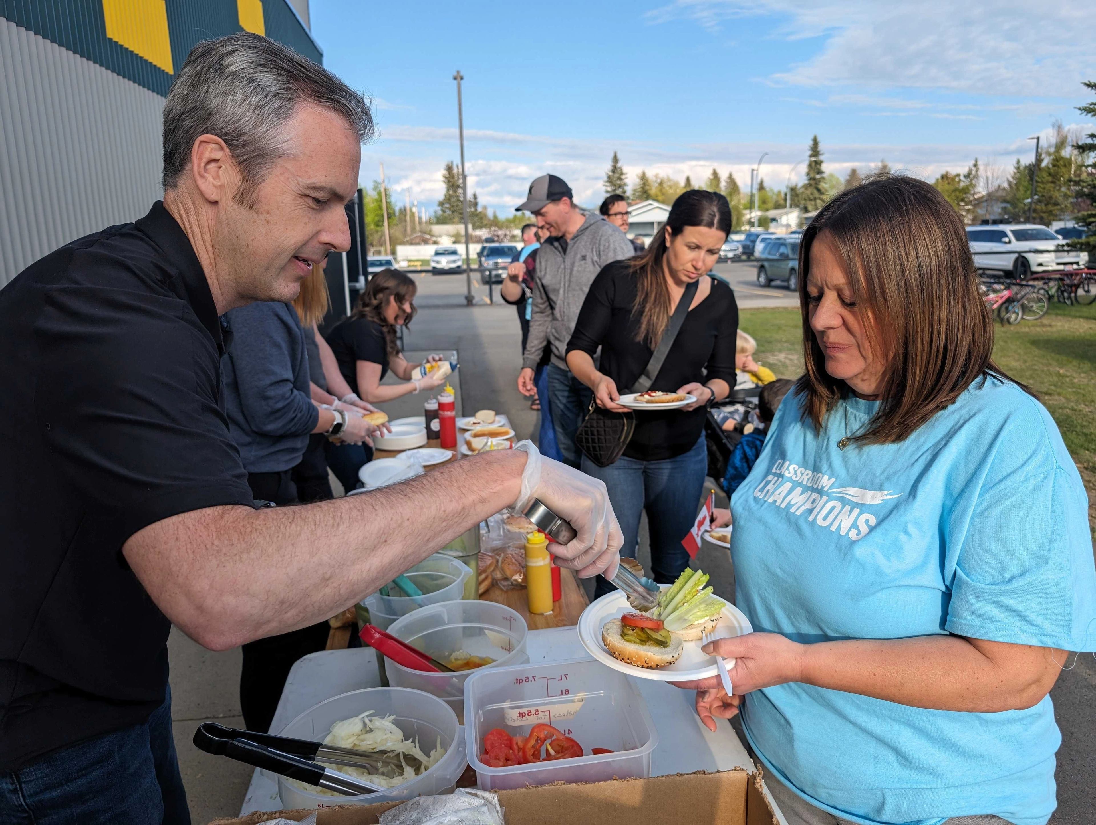 Volunteers serve food to community members wearing Classroom Champions shirts at an outdoor gathering.