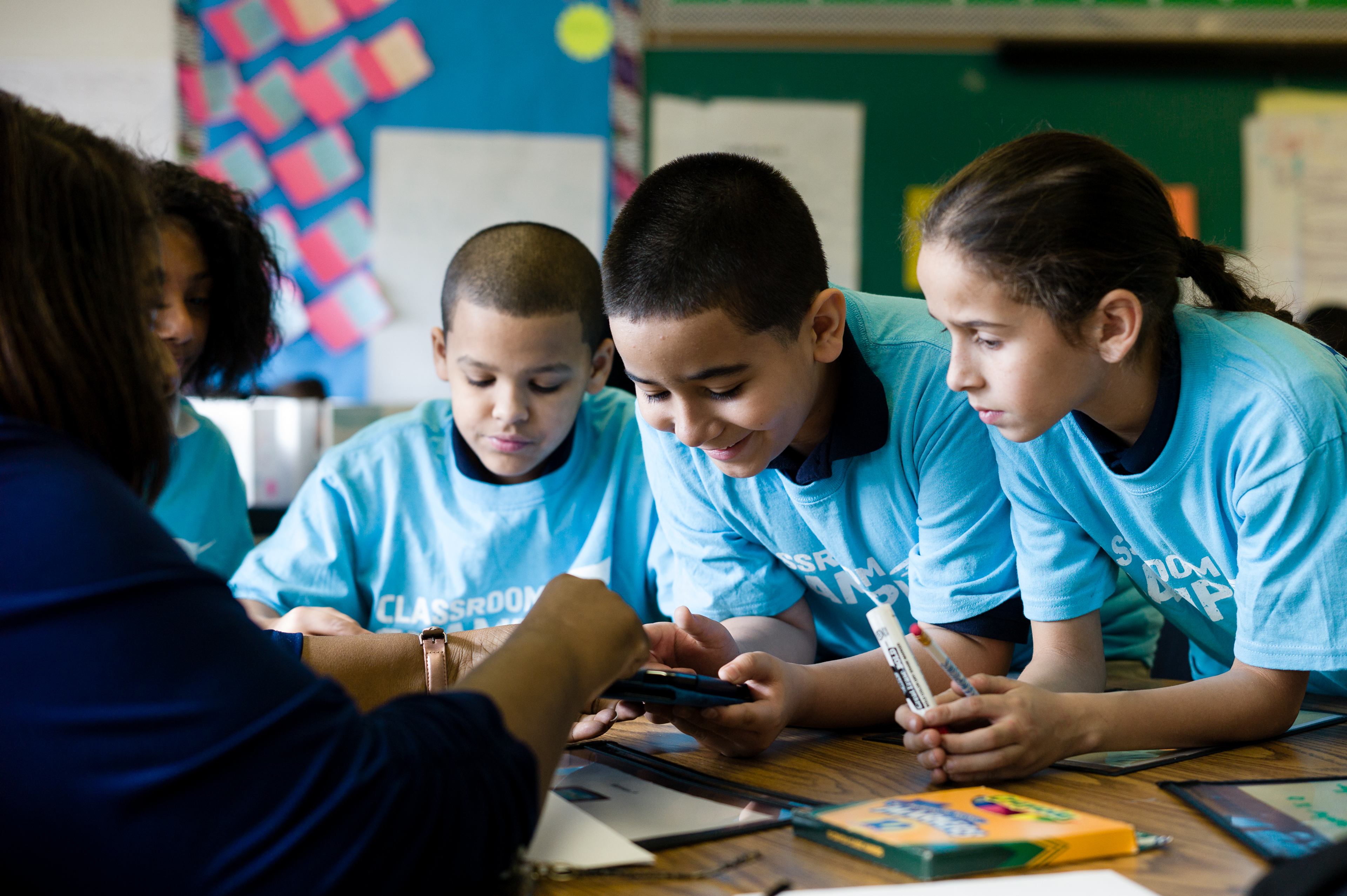 Students wearing Classroom Champions T-shirts gather around a teacher to look at a phone screen.