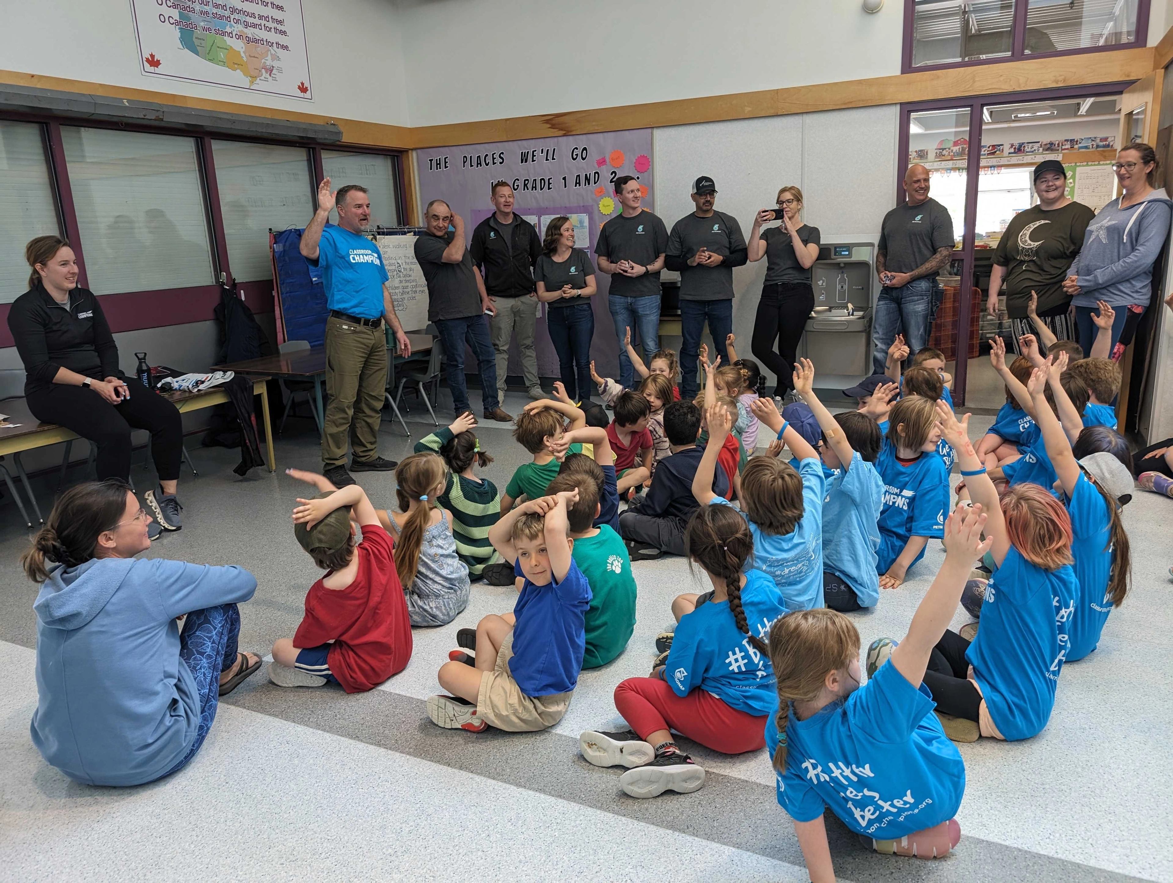 Classroom Champions volunteers stand before a classroom of children raising their hands in Fort St. John.