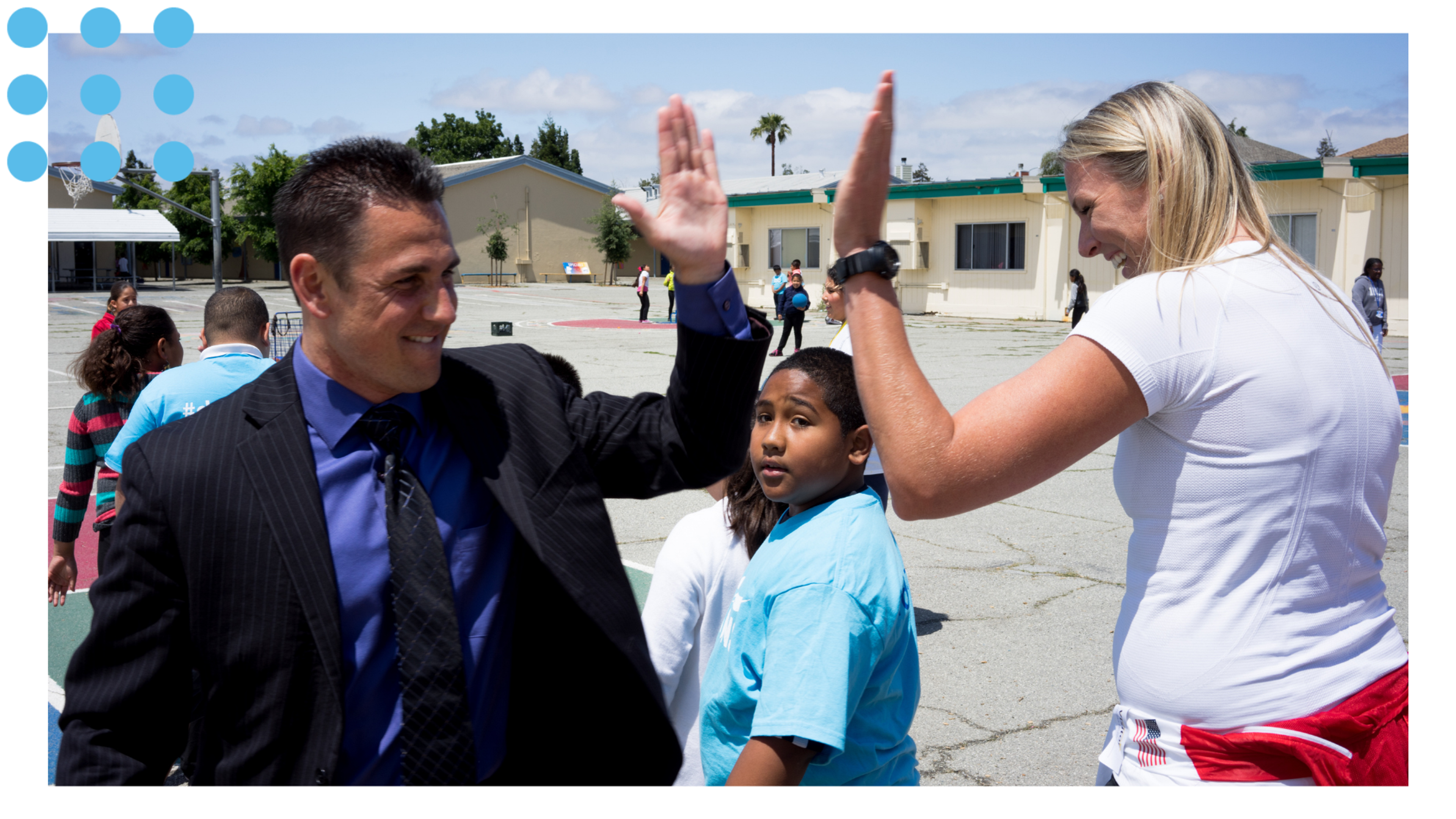 An athlete mentor high-fives a school staff member during a classroom visit on a school playground.