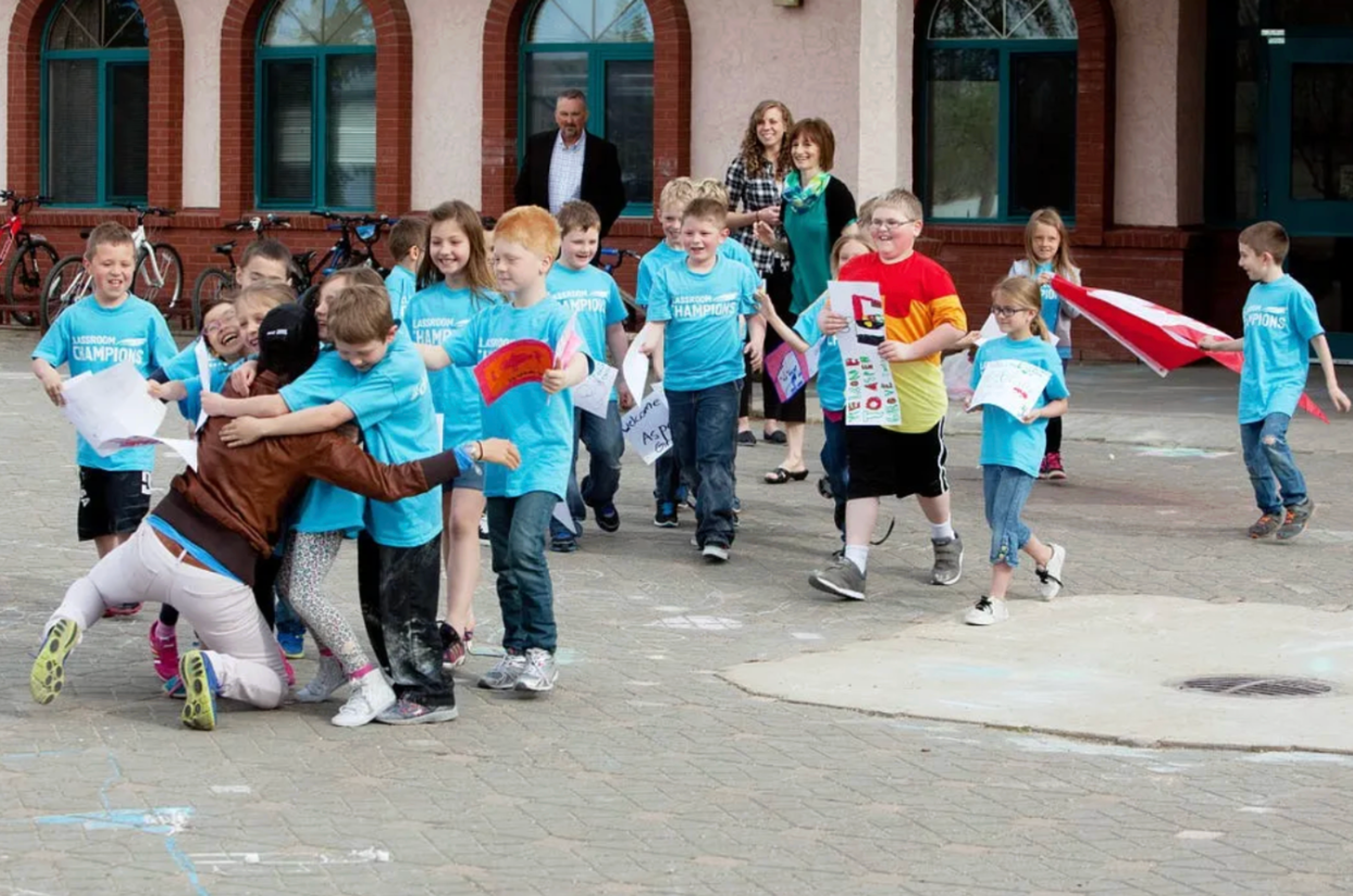 Students in Classroom Champions T-shirts hug a mentor and wave a Canadian flag outside their school.