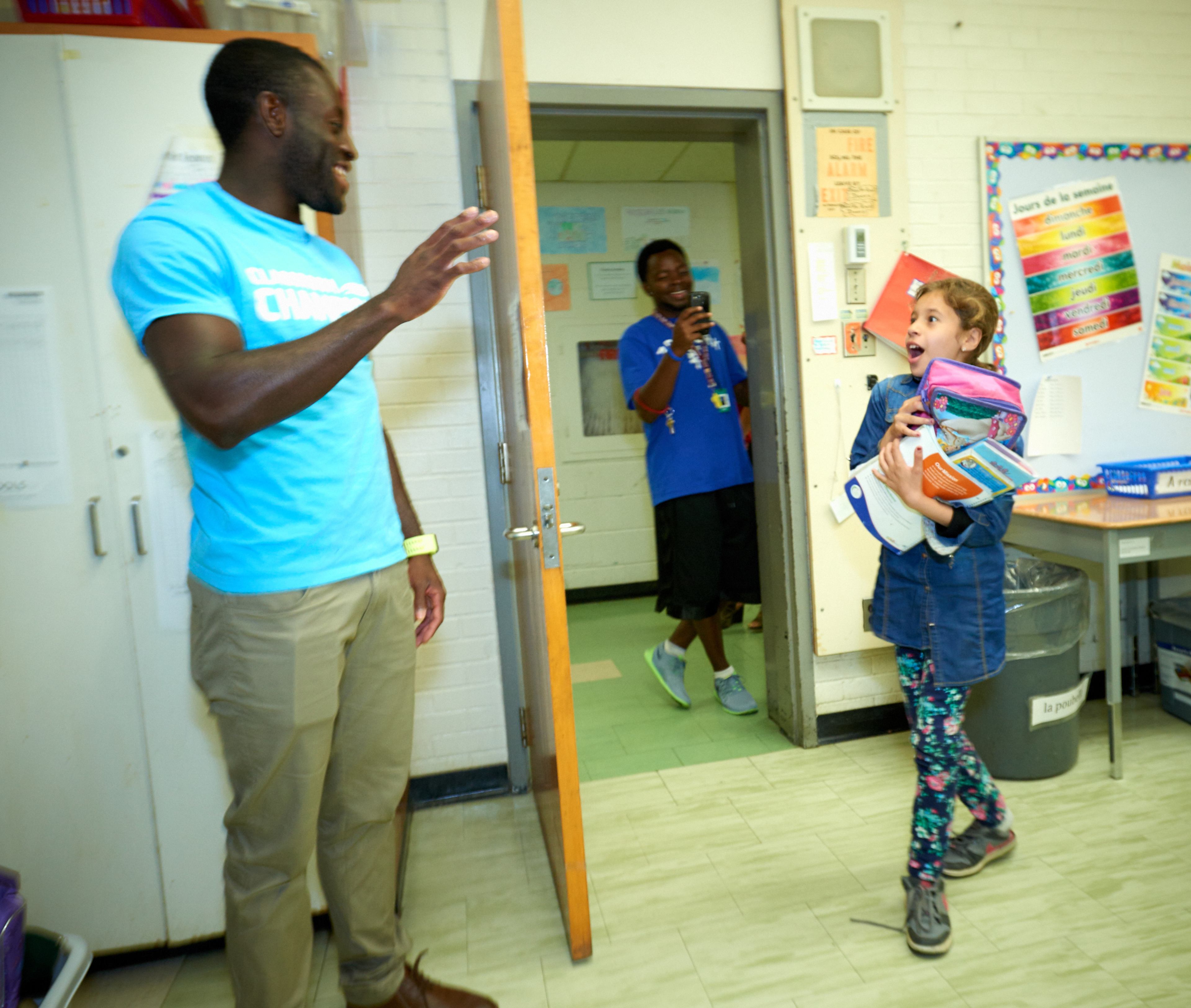 A Classroom Champions volunteer greets a delighted student carrying school supplies in her classroom.