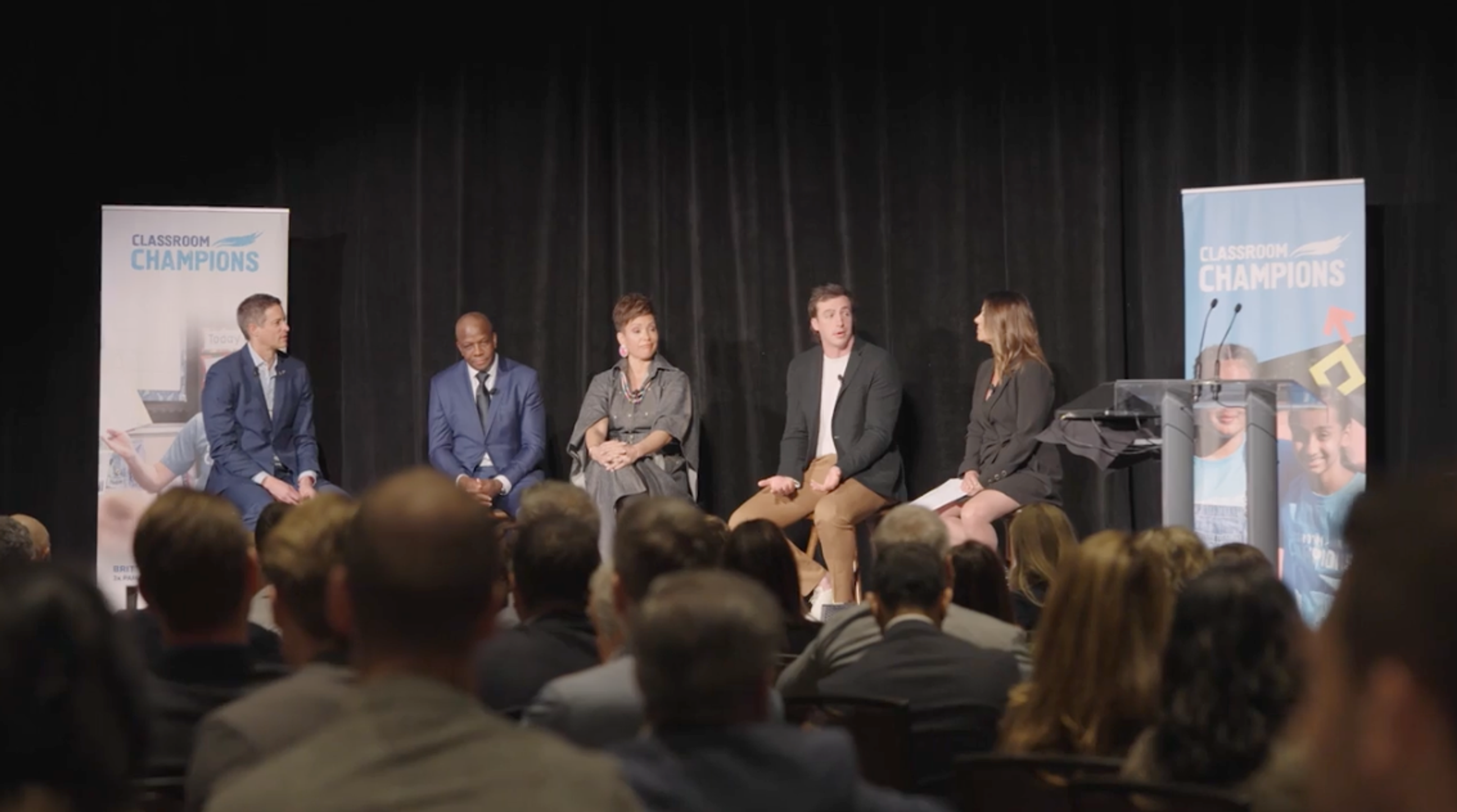 Panelists take part in a discussion on stage at a Classroom Champions event, with an audience seated in front.