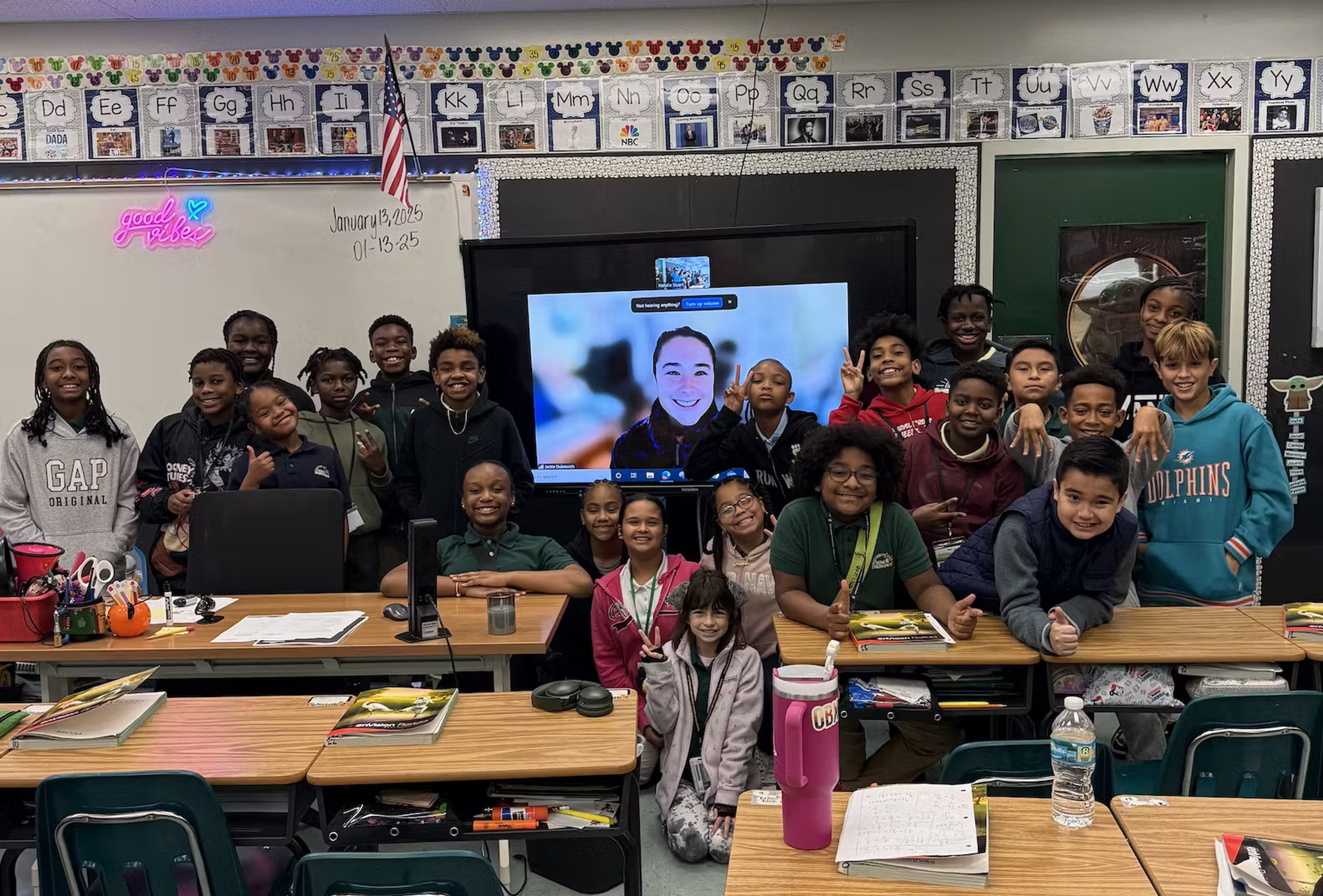 A classroom of students gather smiling around a video call with their mentor displayed on the screen.