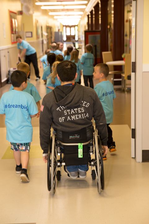 An athlete mentor using a wheelchair walks a school hallway with students wearing #dreambig shirts.