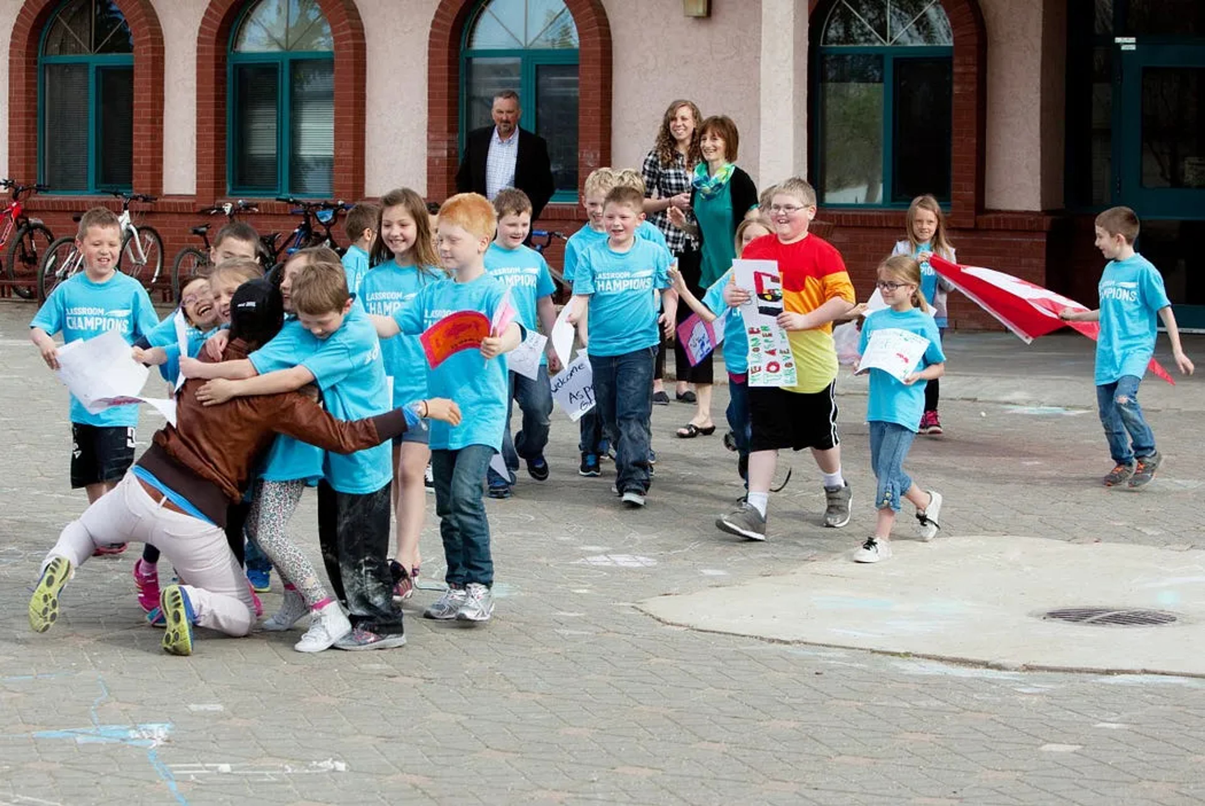 Students in Classroom Champions T-shirts greet a mentor outside their school with signs and a flag.