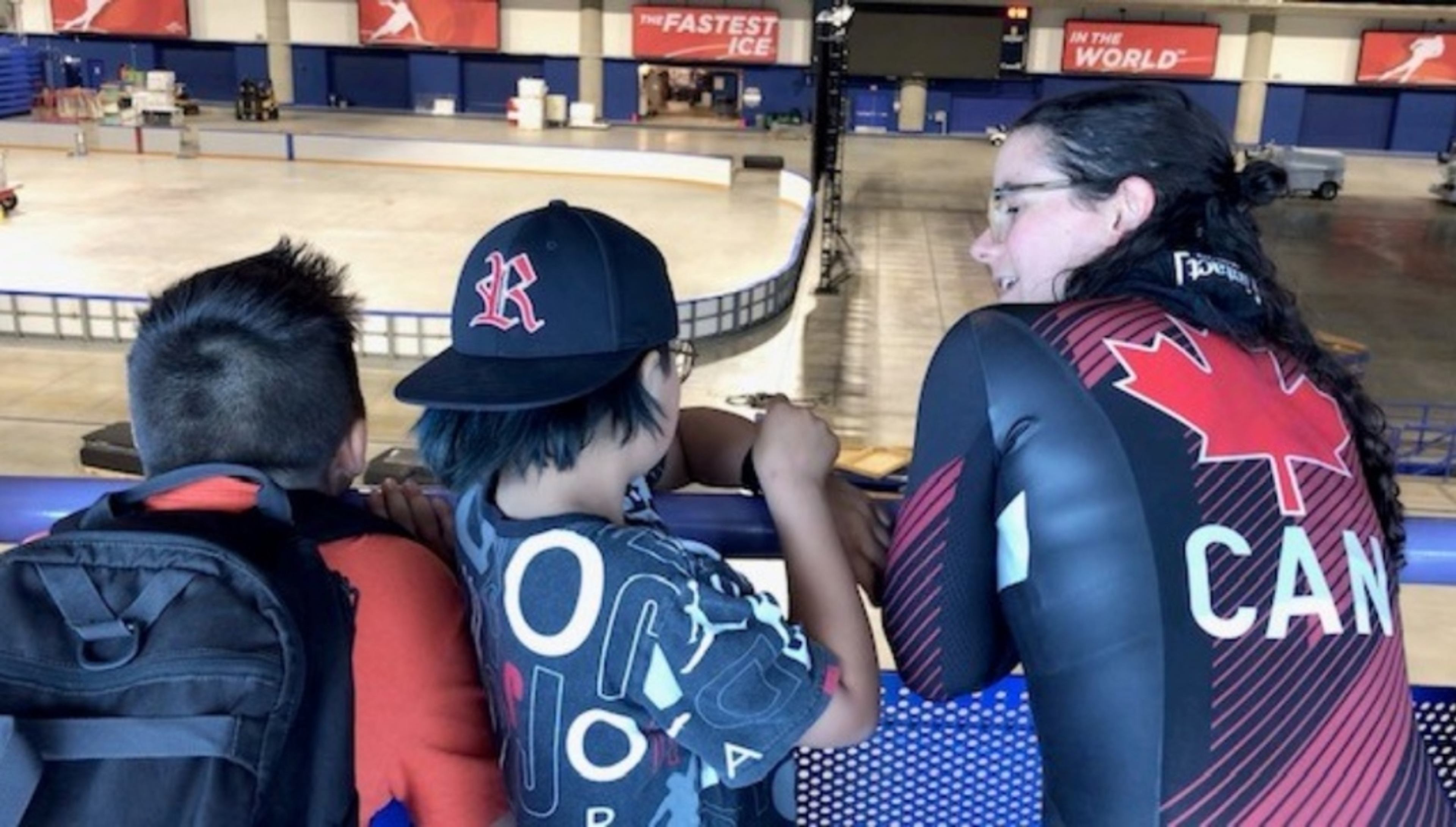 A Team Canada speed-skater in racing gear talks with two students at the edge of an ice rink.