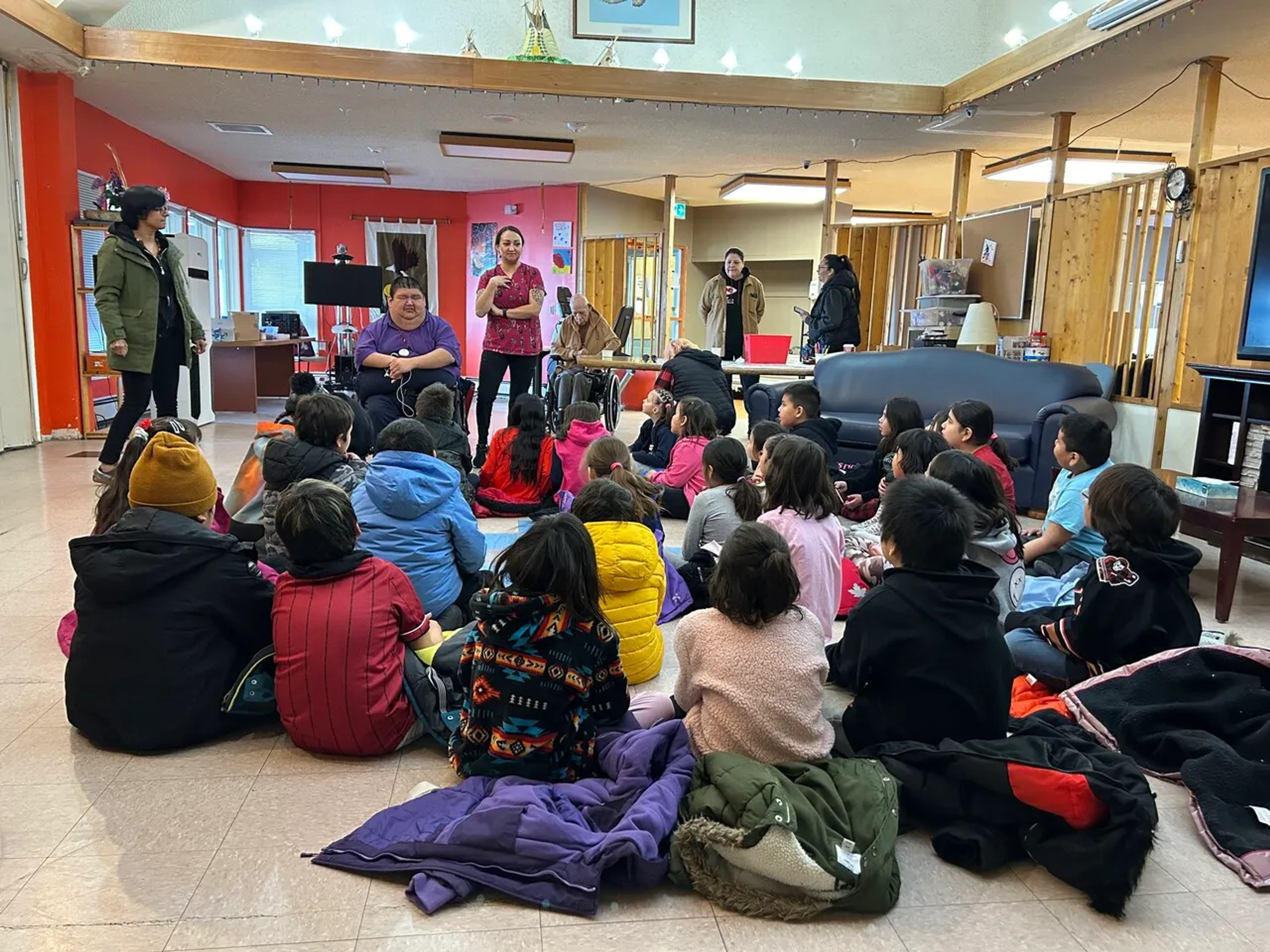 Students sit together on the floor listening to speakers, including a mentor in a wheelchair, in a community room.