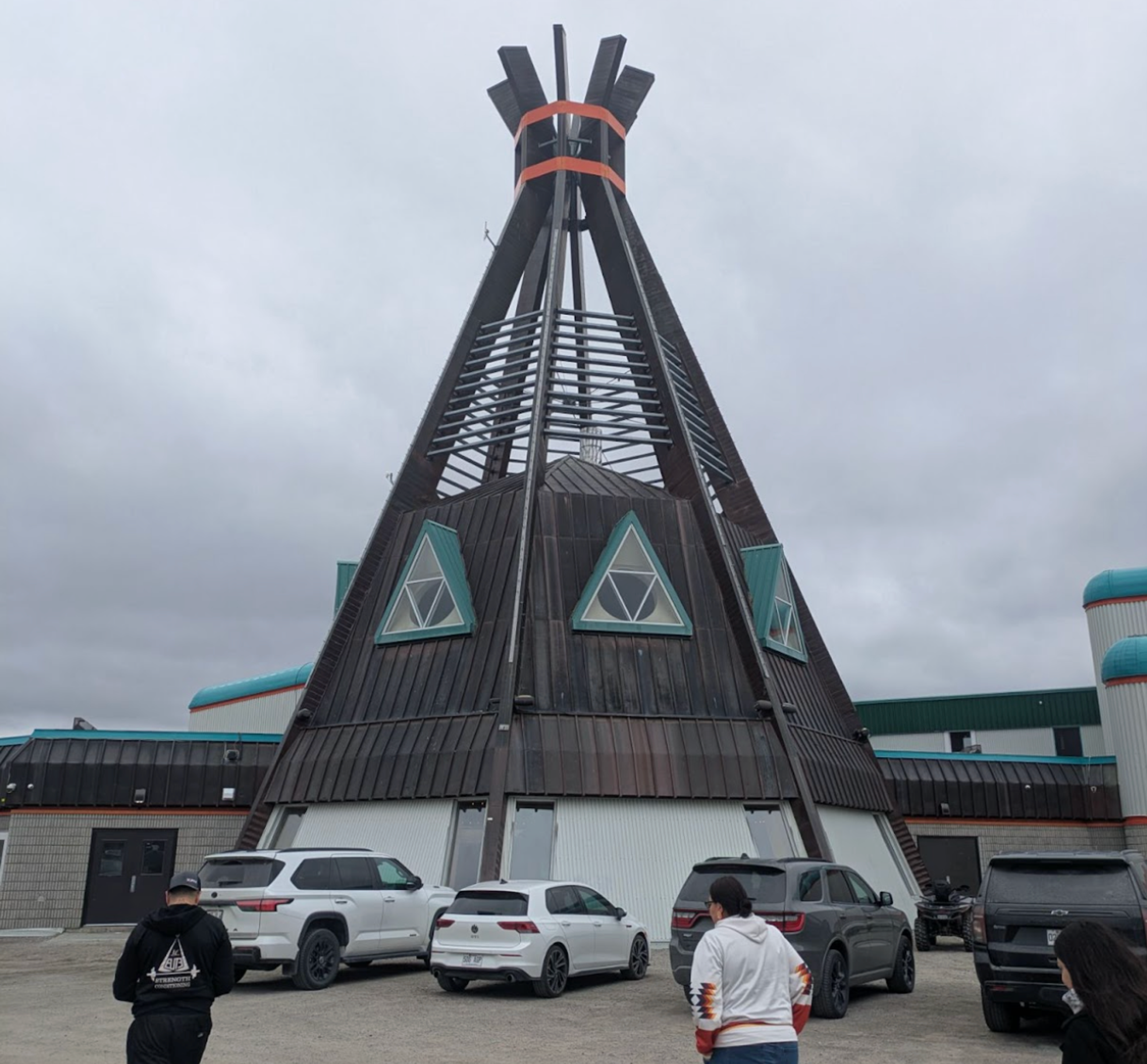 A large teepee-shaped building in Chisasibi, with visitors and parked cars in the foreground.