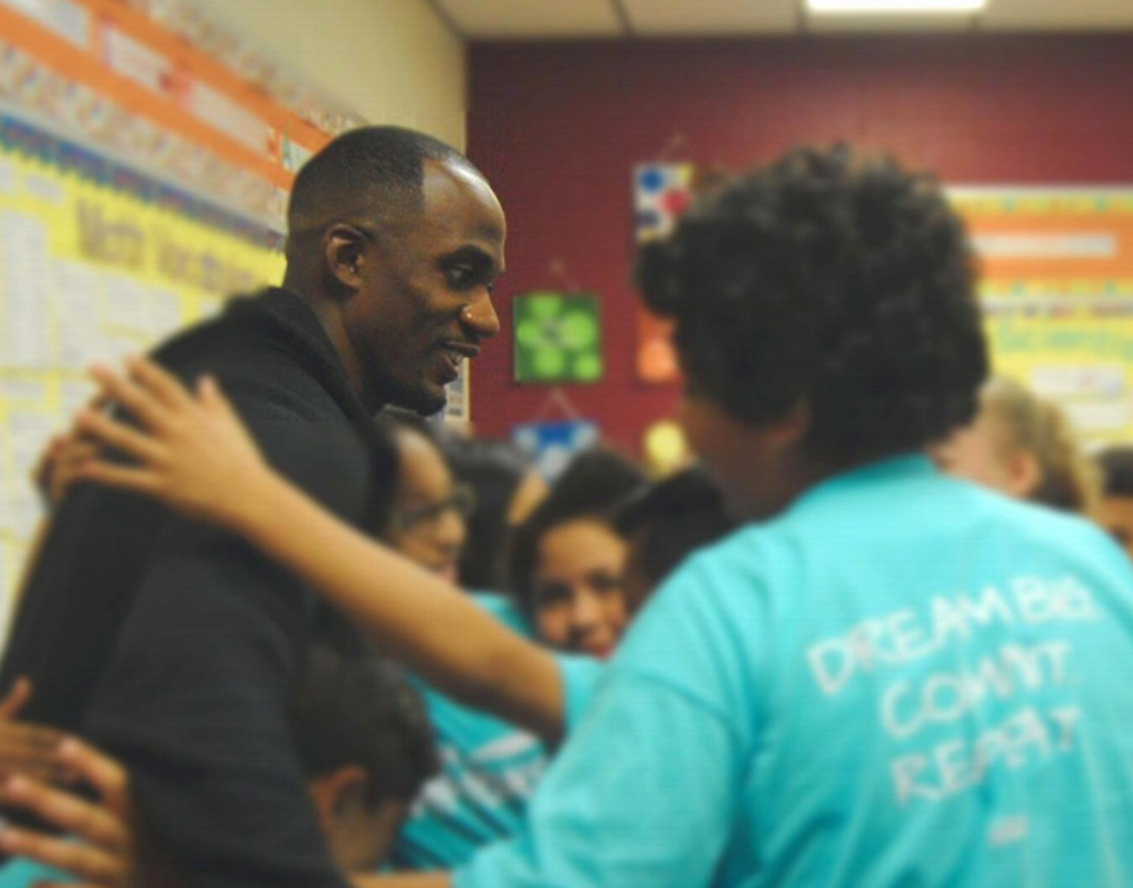 Athlete mentor Lex Gillette hugs a student wearing a shirt reading "Dream Big, Commit, Repeat" in a classroom.