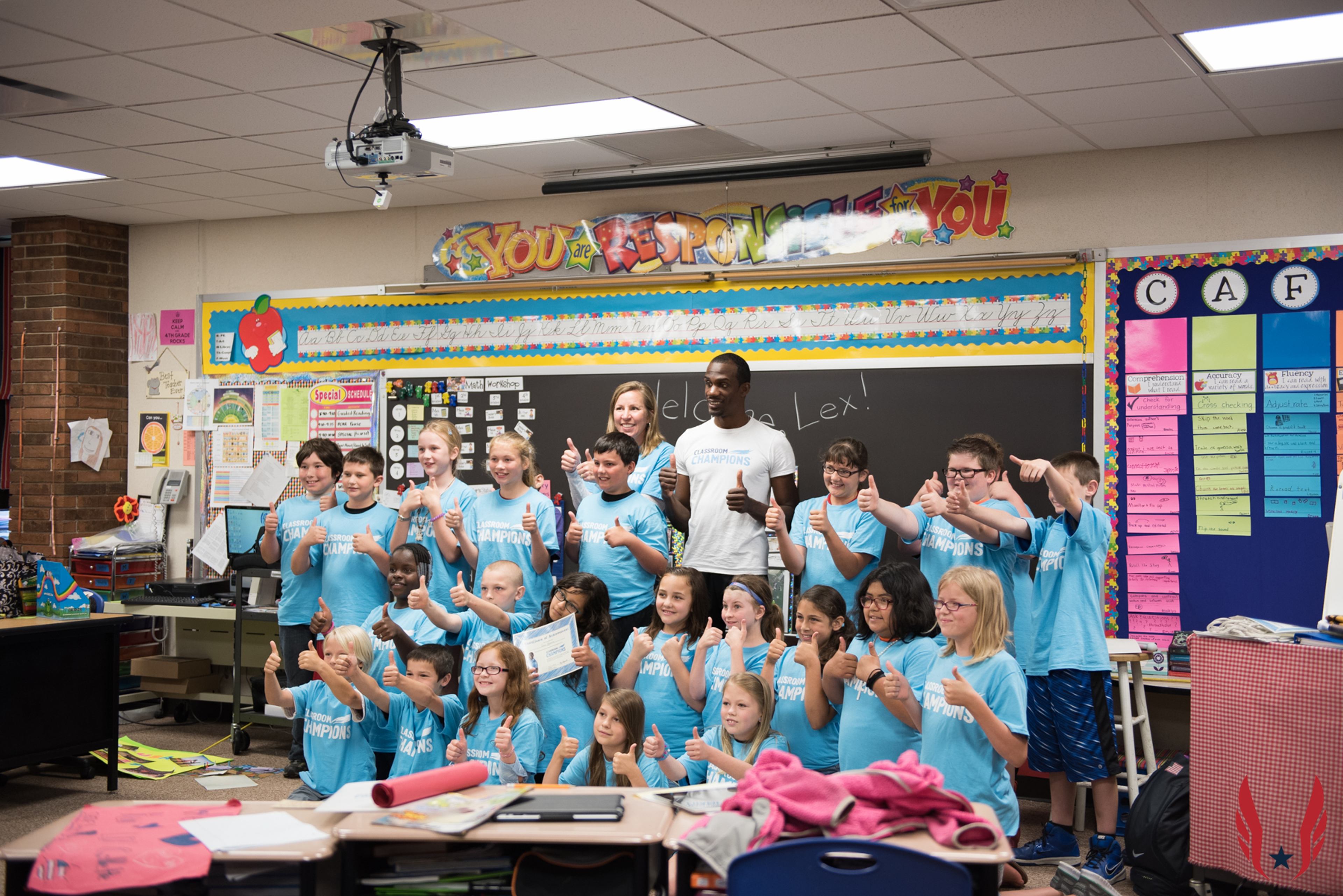 A classroom of students in matching Classroom Champions shirts give thumbs up alongside their mentor and teacher.