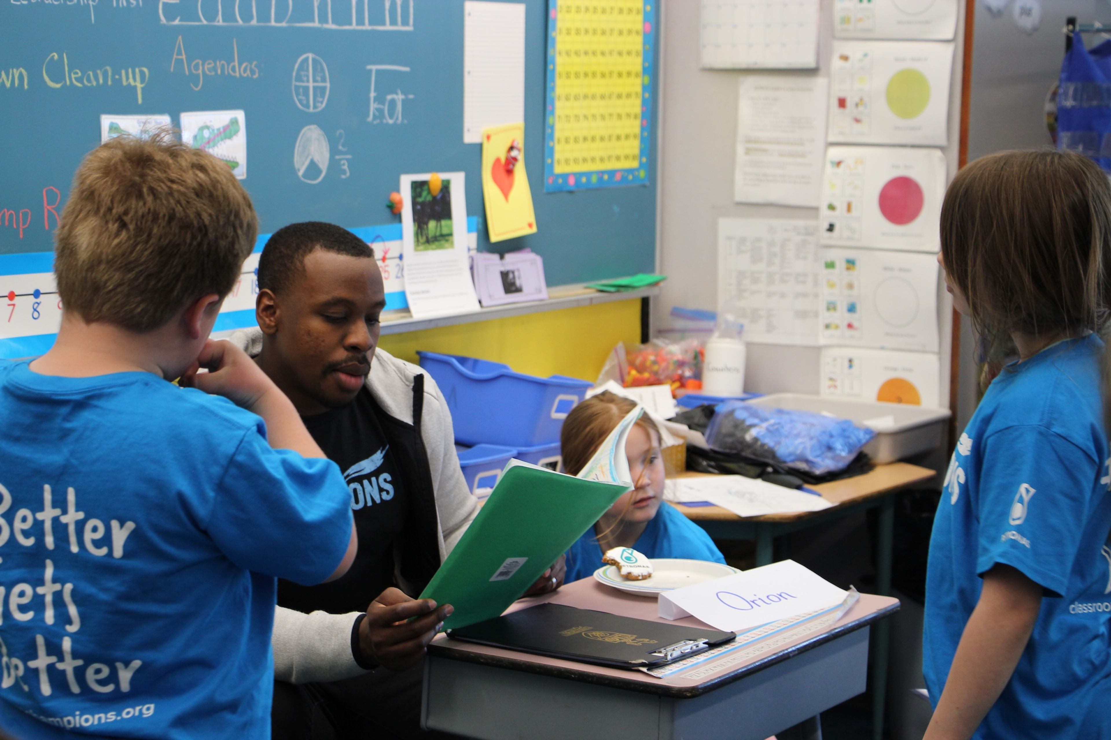 An athlete mentor reviews a folder with students wearing Classroom Champions shirts in a classroom.