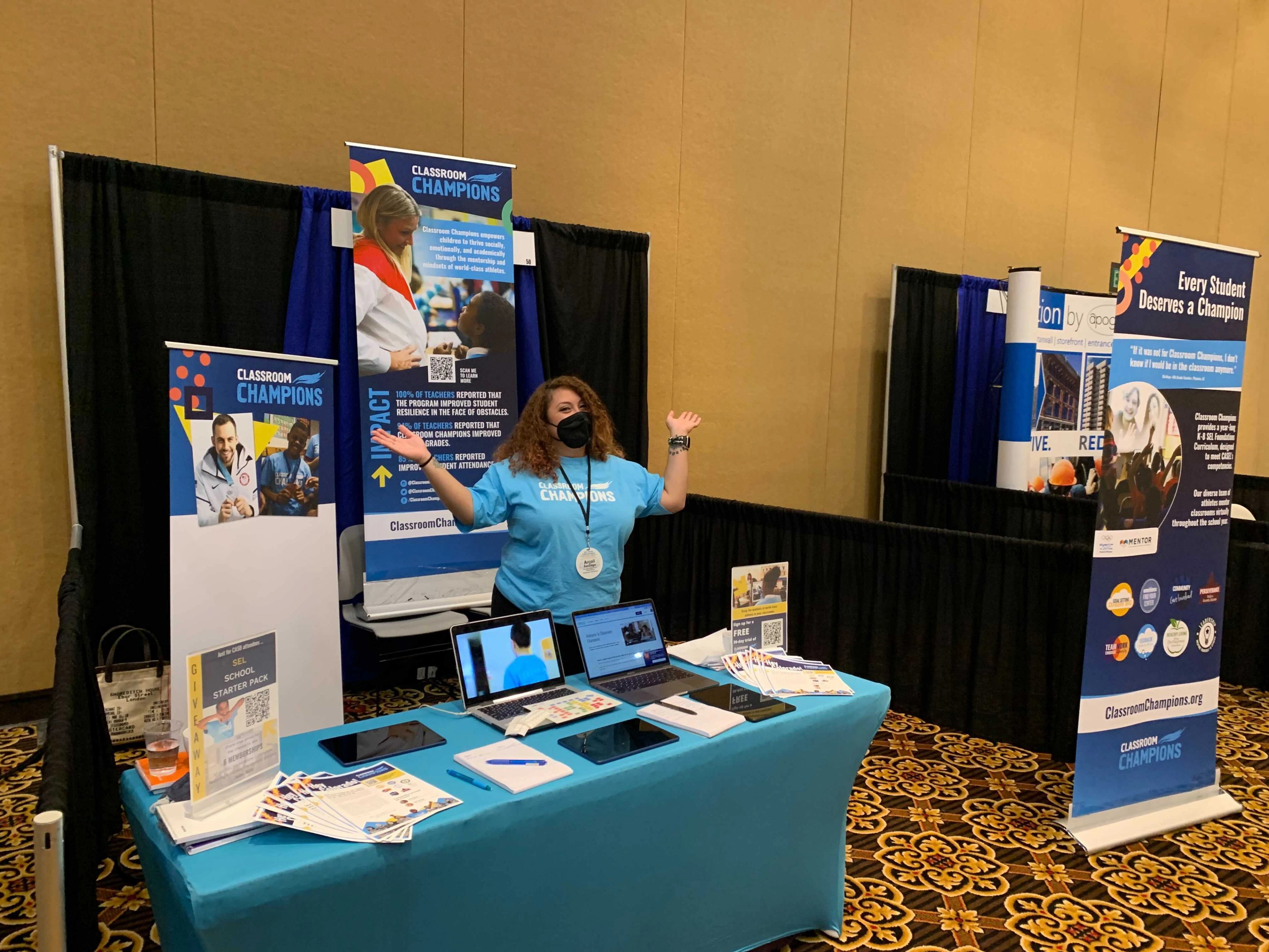 A Classroom Champions staff member greets visitors at a conference booth featuring program banners and materials.