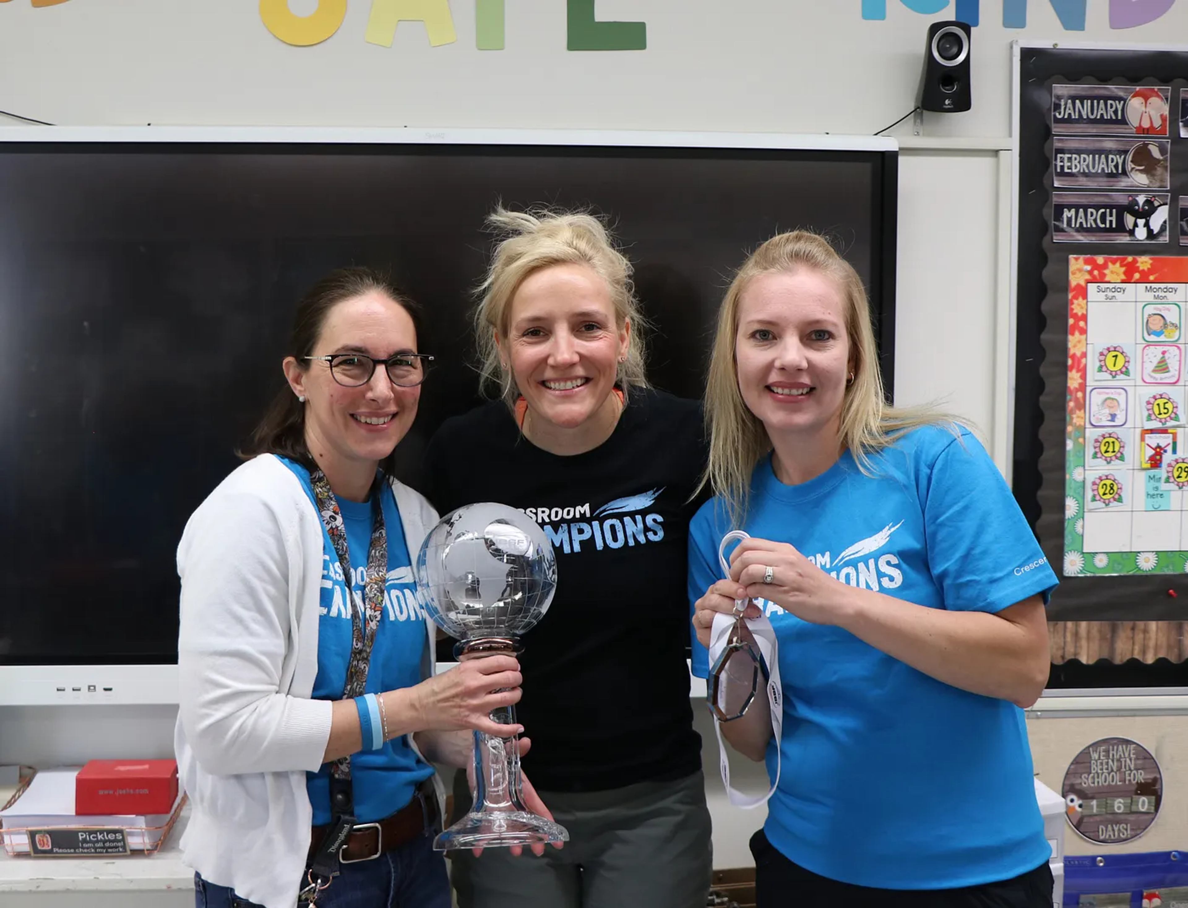 An athlete mentor poses with two teachers holding a trophy in a classroom during a Classroom Champions visit.