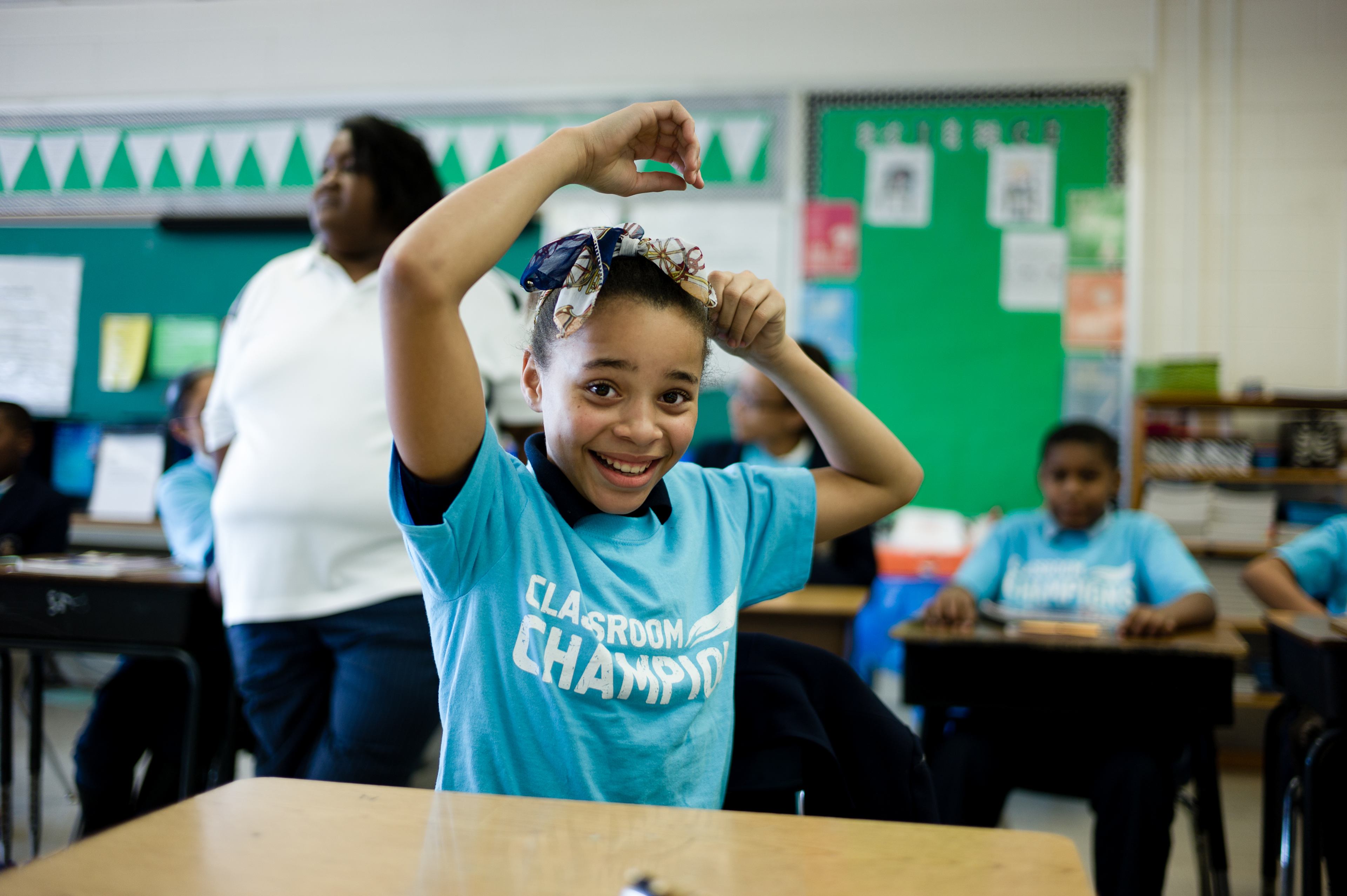 A smiling student wearing a Classroom Champions T-shirt poses at her desk during class.