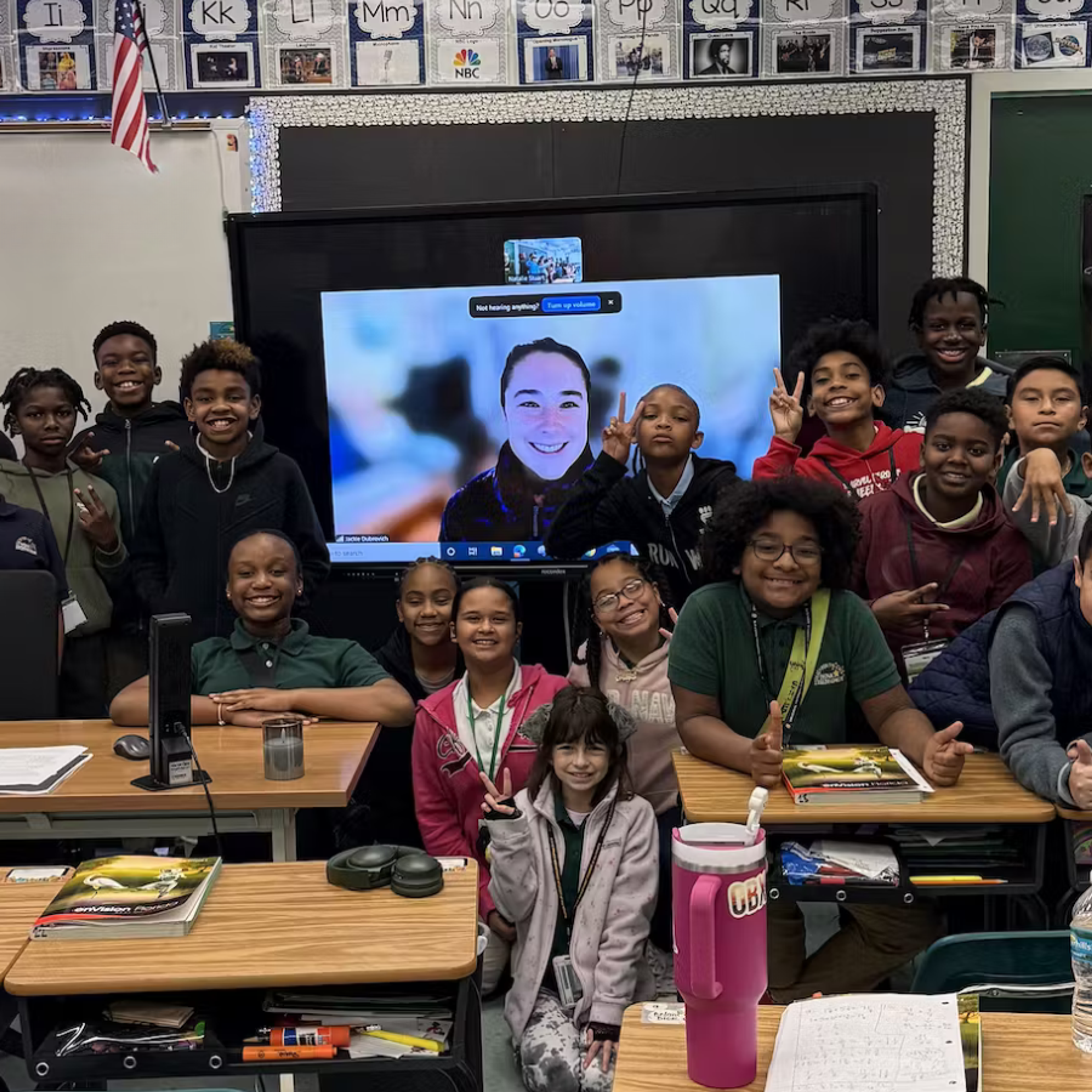 A classroom of students pose together during a video call with their mentor on screen.