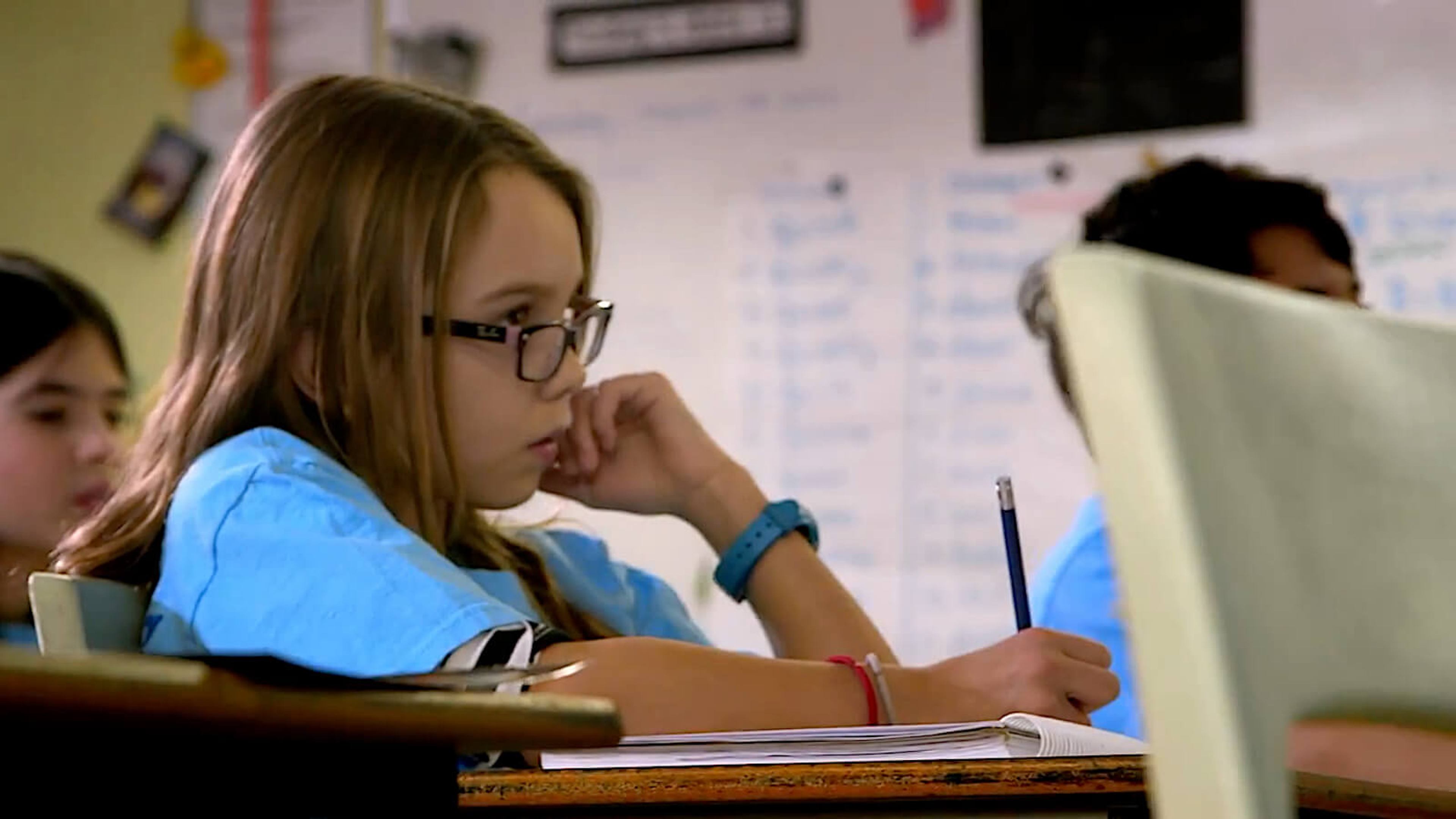 A girl in a blue t-shirt writes in a notebook in class.