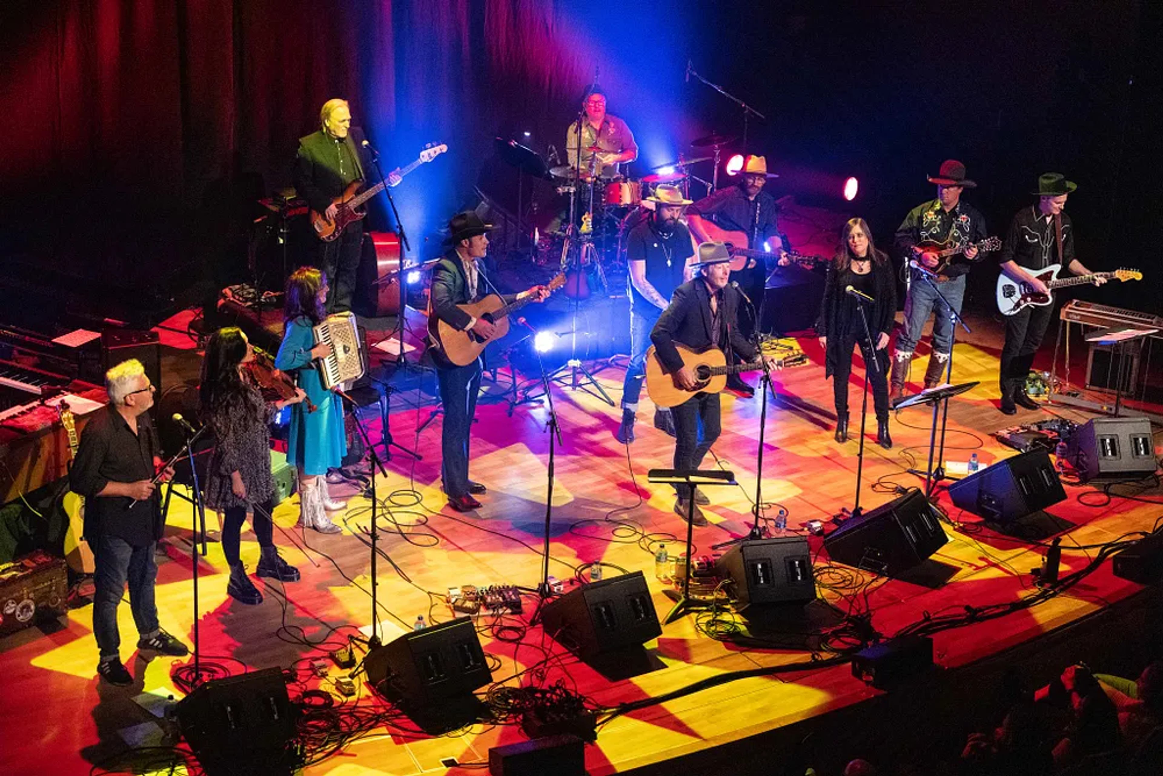 A large band performs on stage with guitars, fiddle, accordion, and colorful lighting at a Classroom Champions benefit concert.
