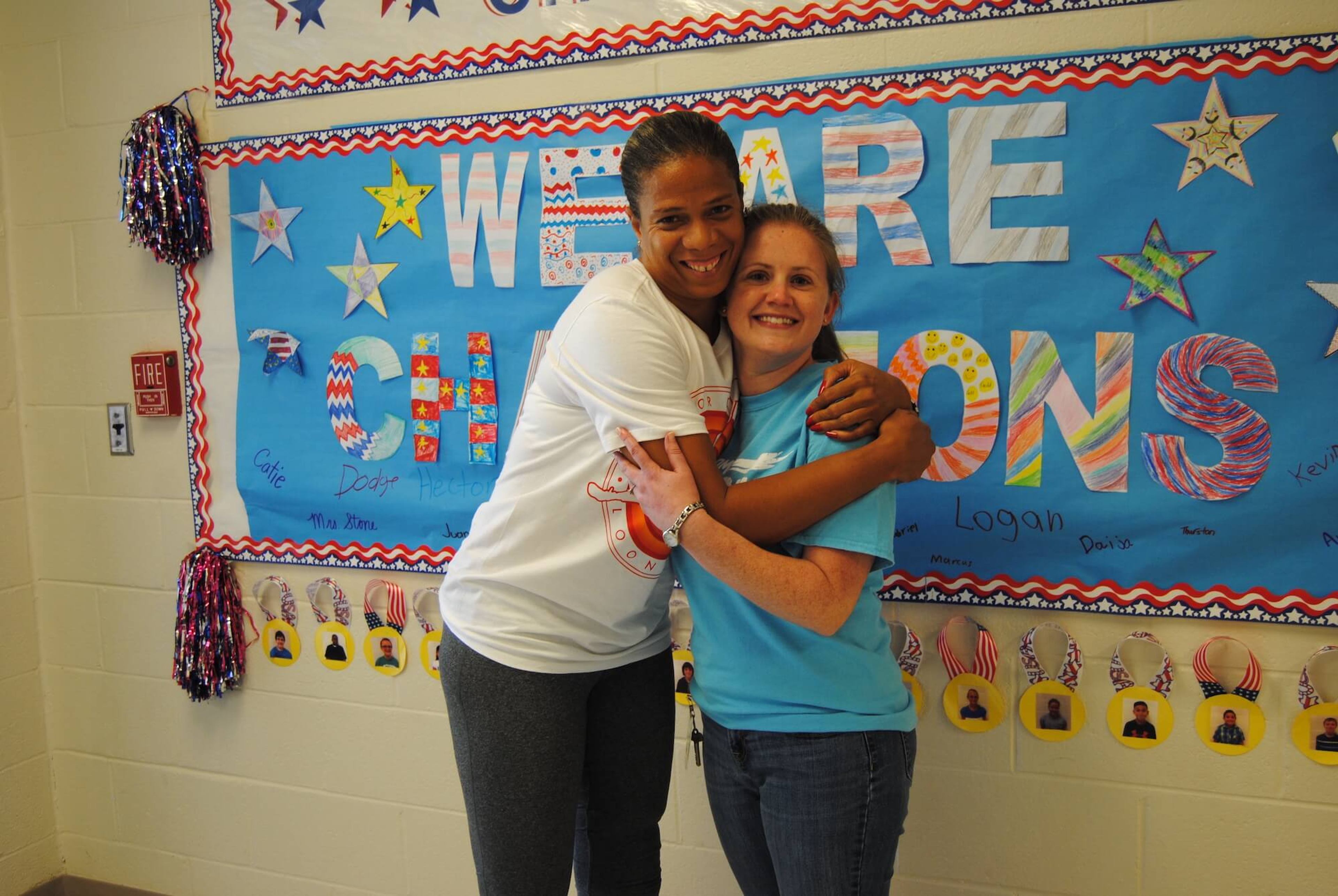 Two women hugging in front of a sign reading 'We Are Champions'