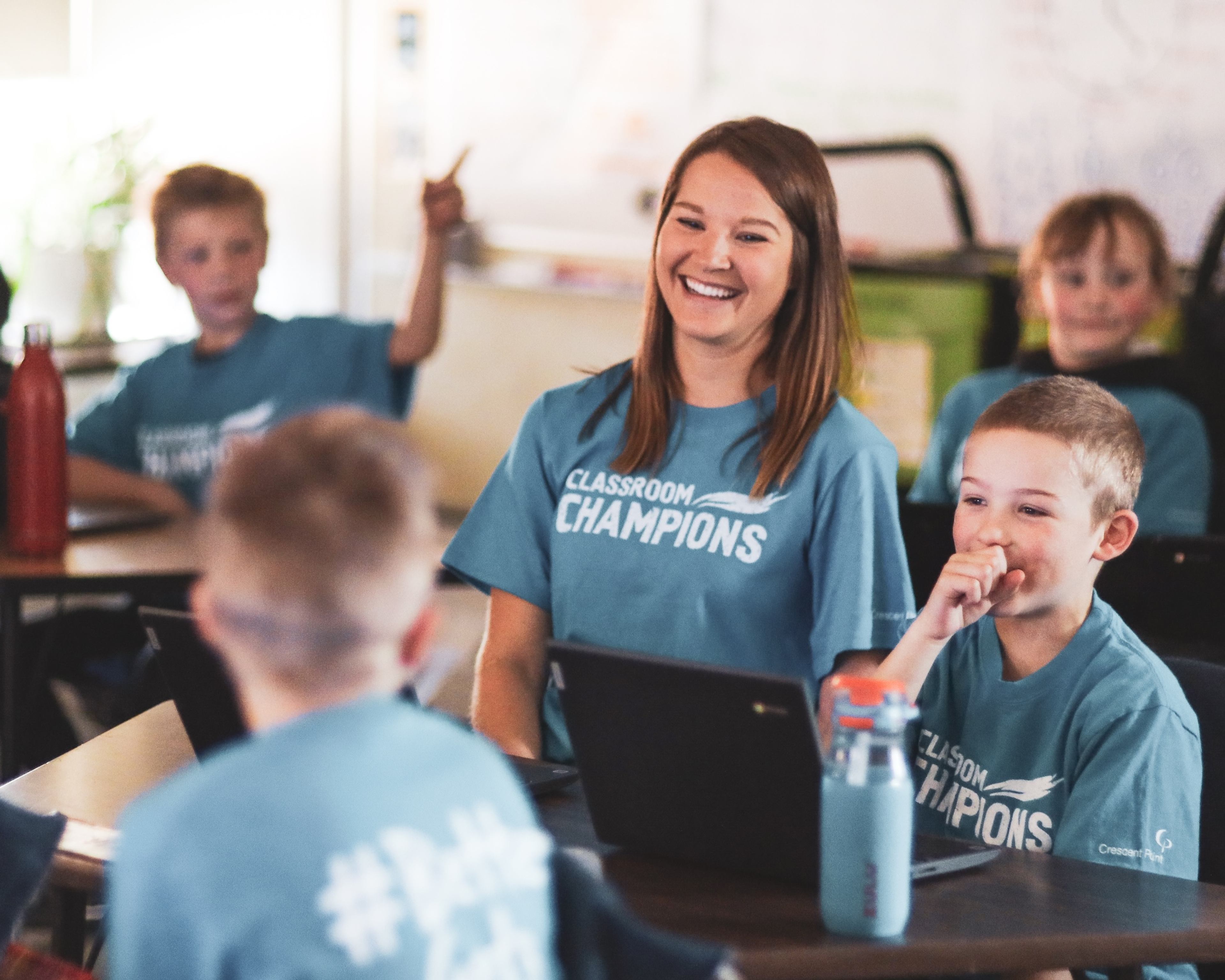 A teacher and students in Classroom Champions t-shirts smile together during a classroom activity with laptops.
