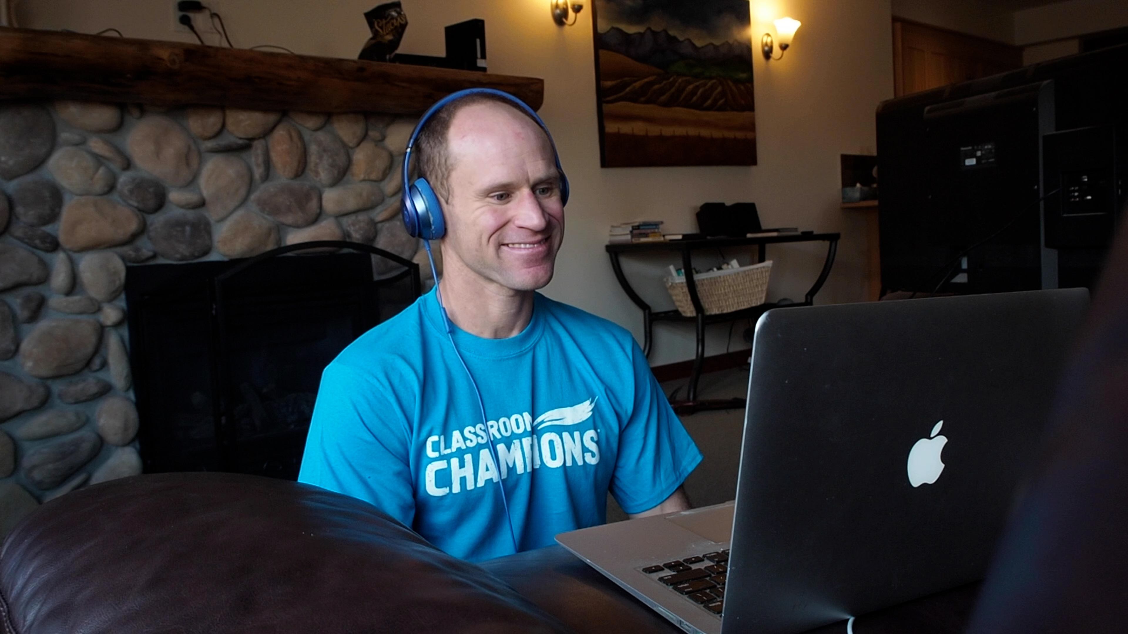 Dan Cnossen, wearing a Classroom Champions t-shirt, smiles during a virtual classroom mentoring session on his laptop.