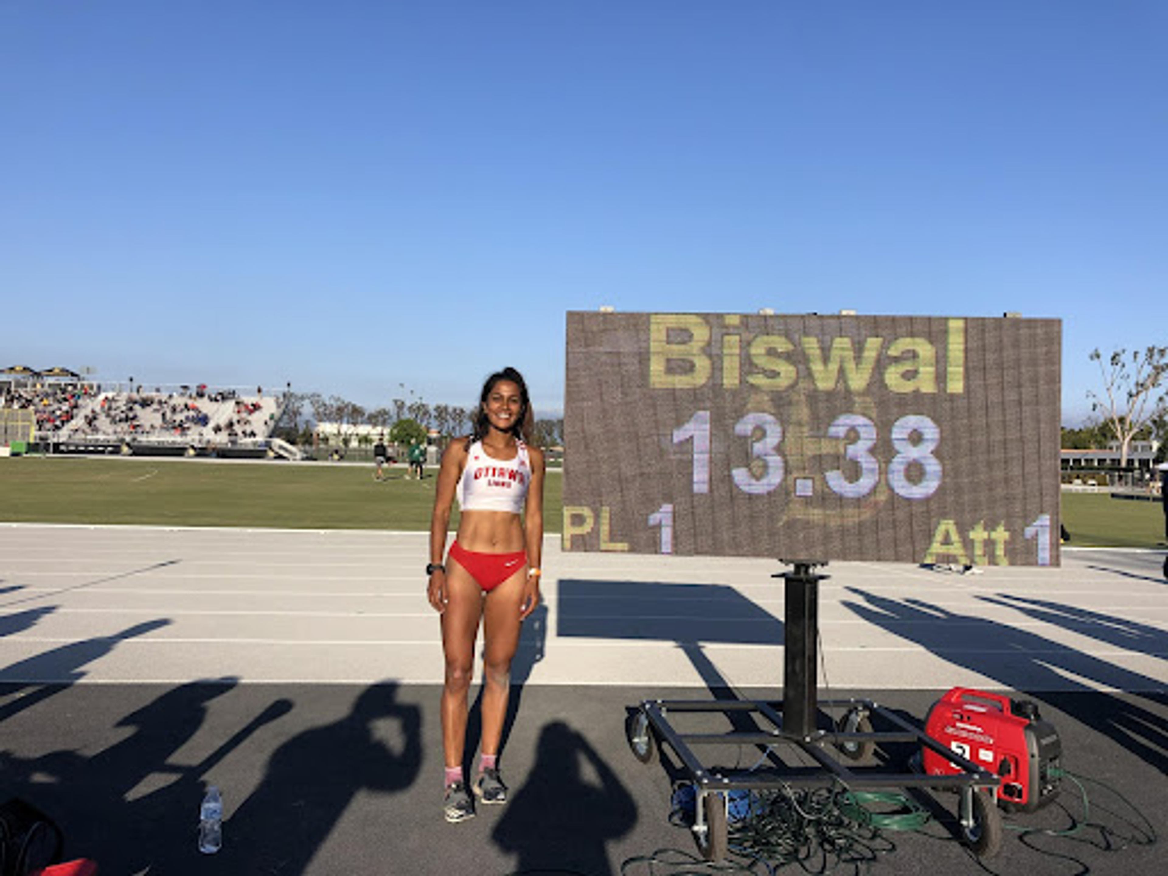 Athlete Divya Biswal stands by a scoreboard showing her first-place 13.38 result at a track meet.