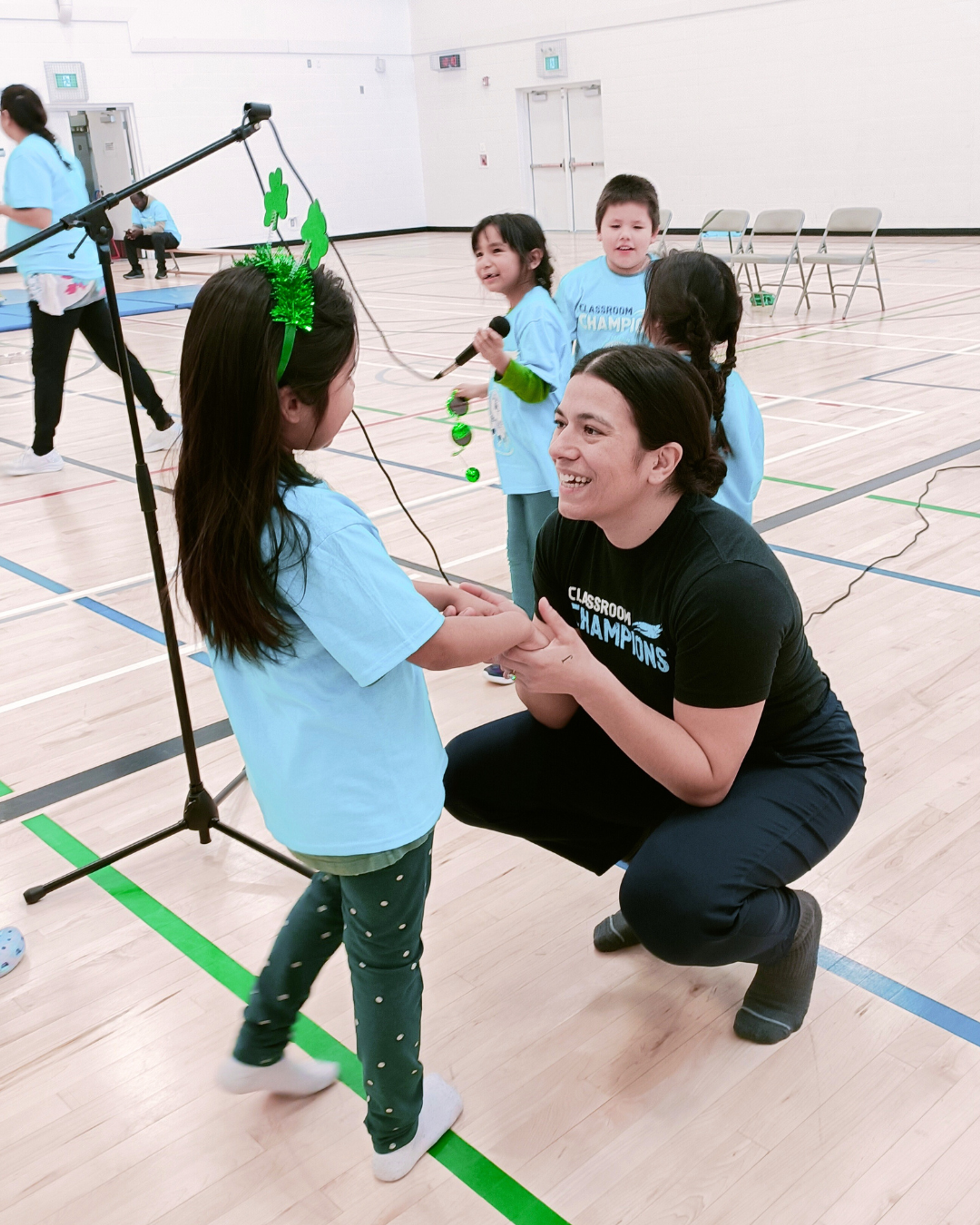 A Classroom Champions mentor kneels smiling with a student in a gymnasium during a school event.