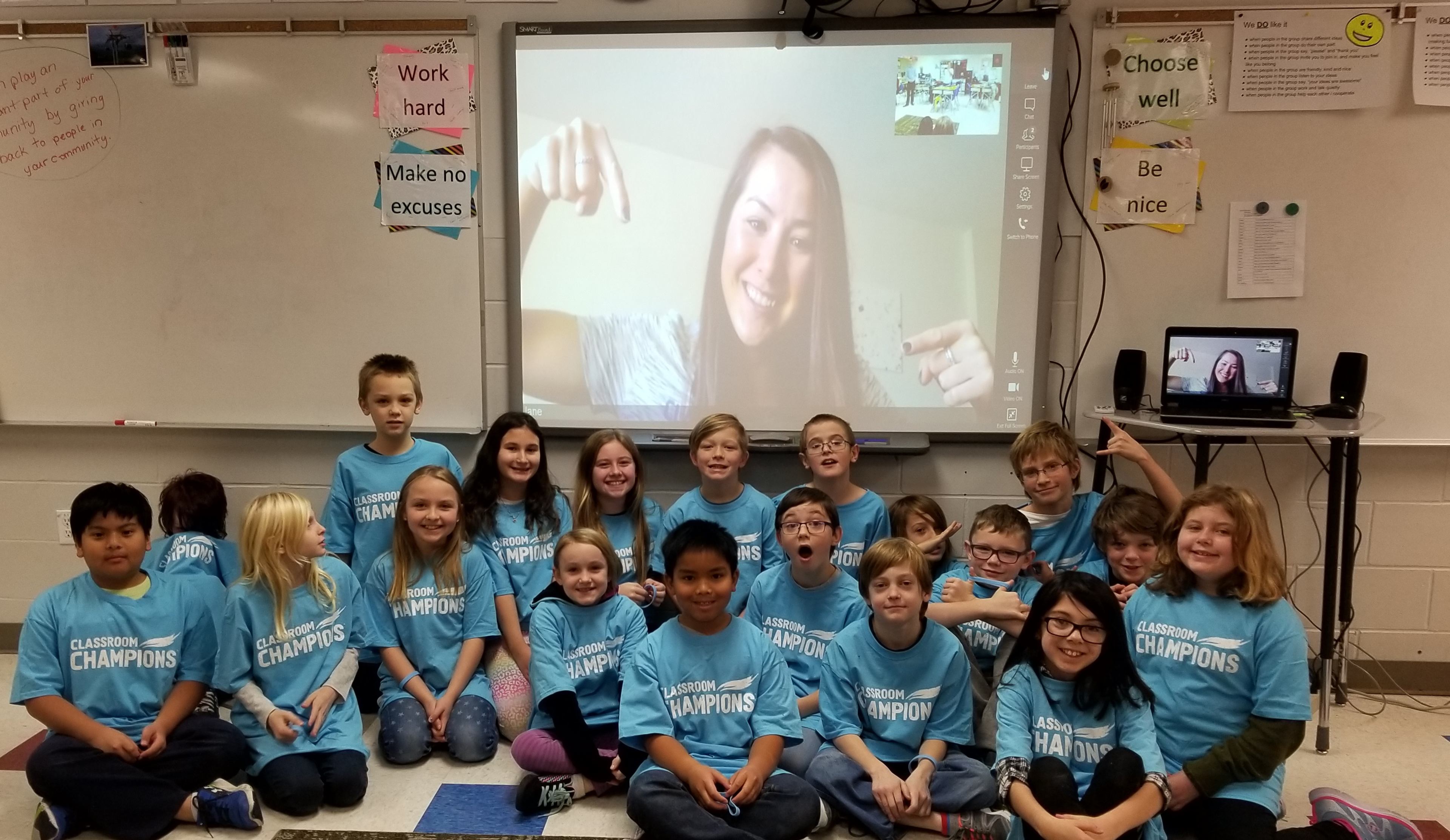 Students wearing Classroom Champions shirts pose for a video call with their athlete mentor on a classroom screen.
