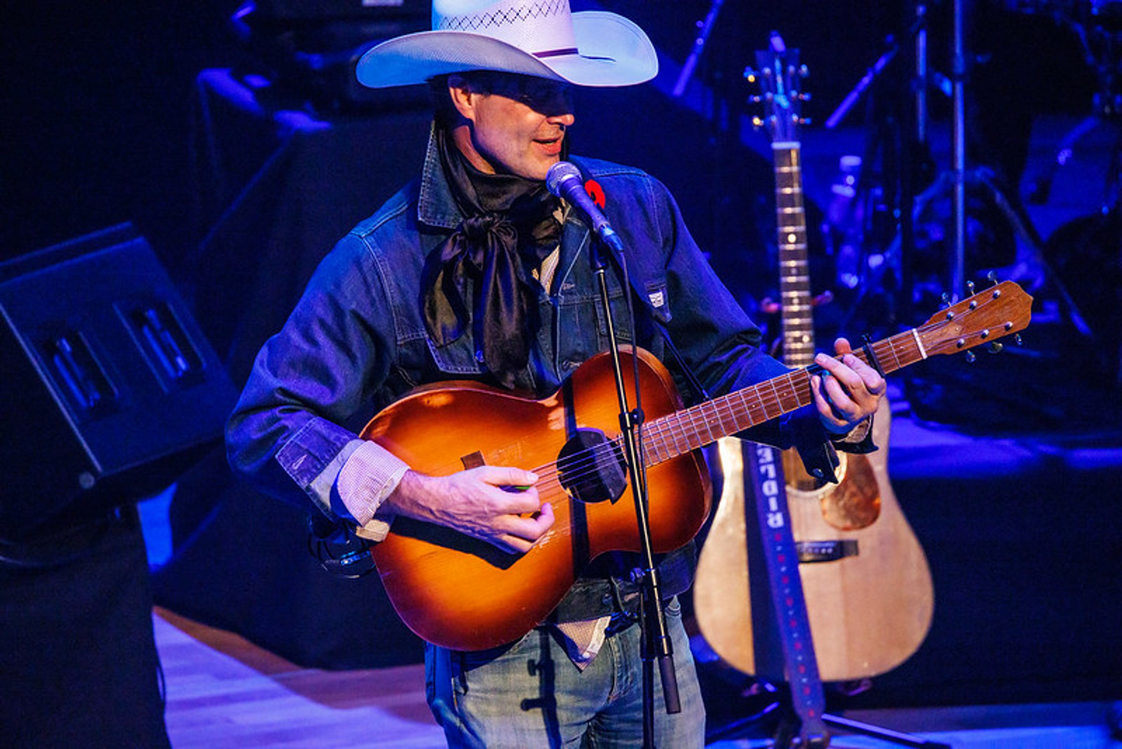 A musician in a cowboy hat performs onstage with an acoustic guitar at a Concert for Classrooms event.
