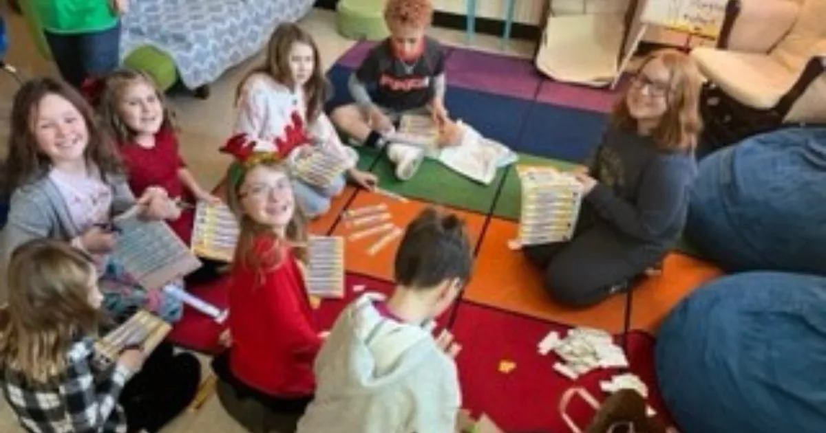 Classroom students sit together on a colorful rug working on a group service learning craft project.