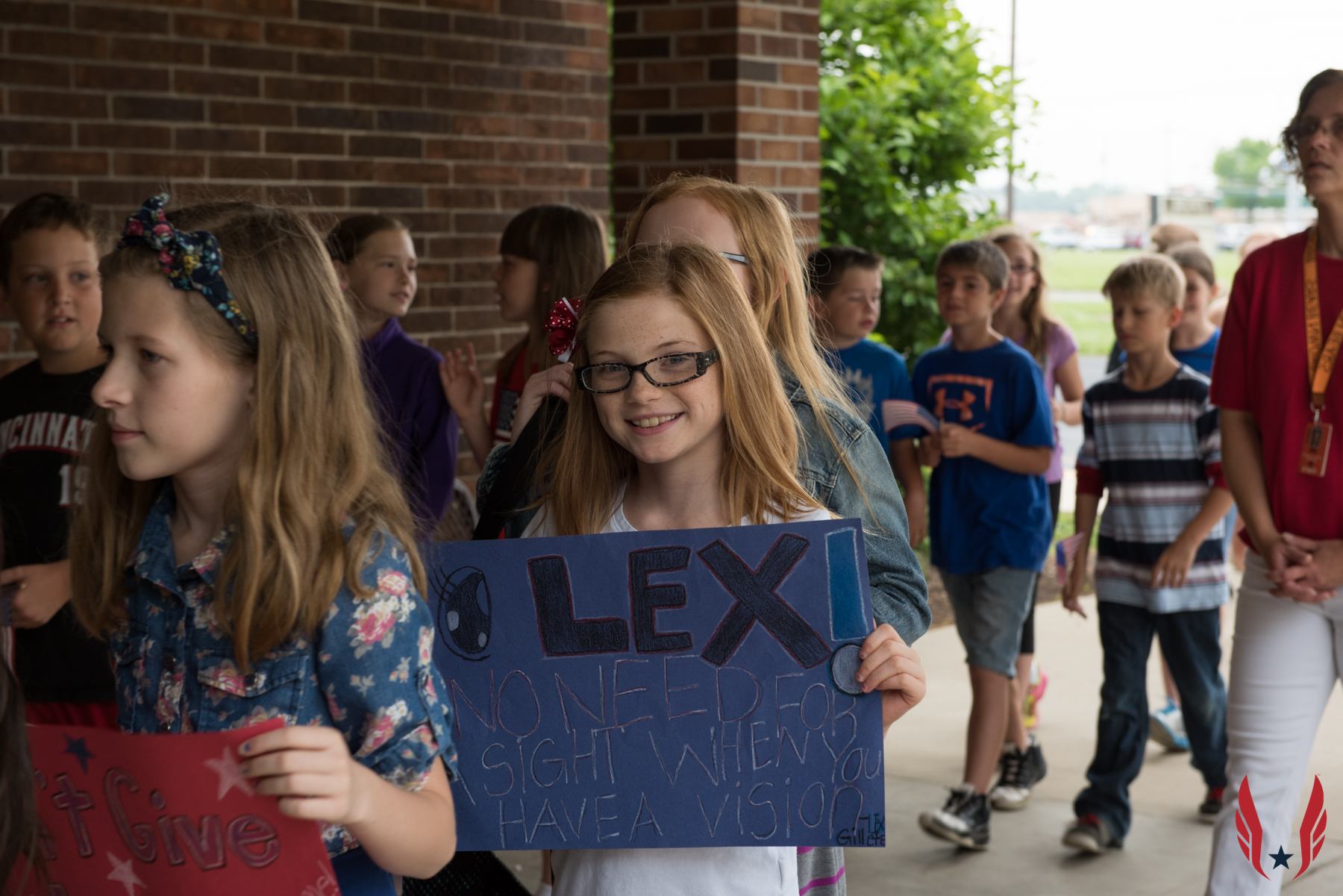 A student holds a handmade sign for mentor Lex reading "No need for sight when you have a vision" as classmates walk outside.