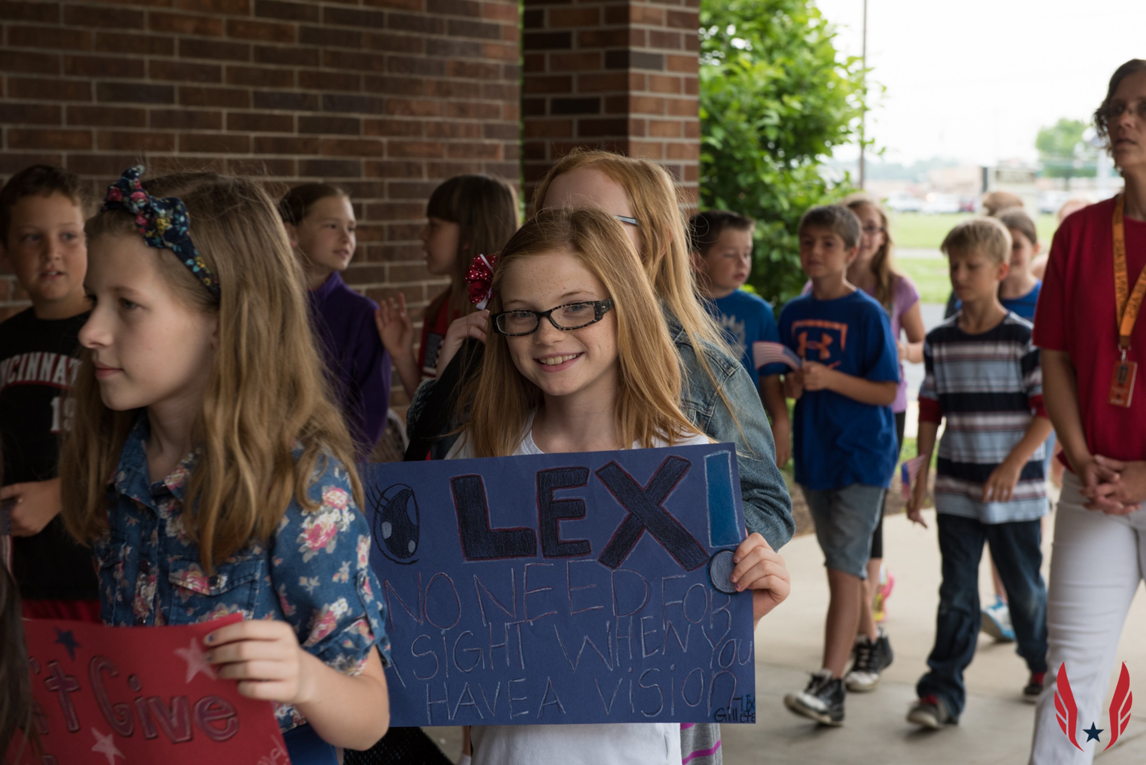 A student holds a handmade sign for mentor Lex reading "No need for sight when you have a vision" as classmates walk outside.