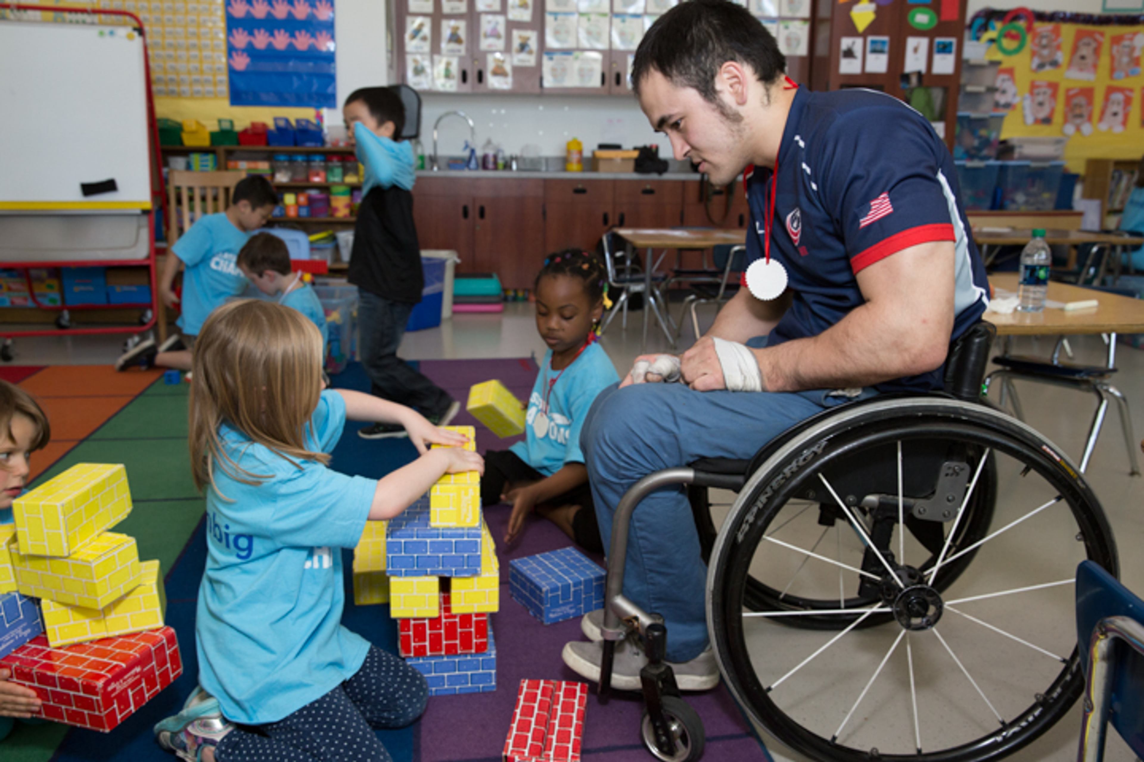 An athlete mentor using a wheelchair builds a block tower on the floor with young students in a classroom.