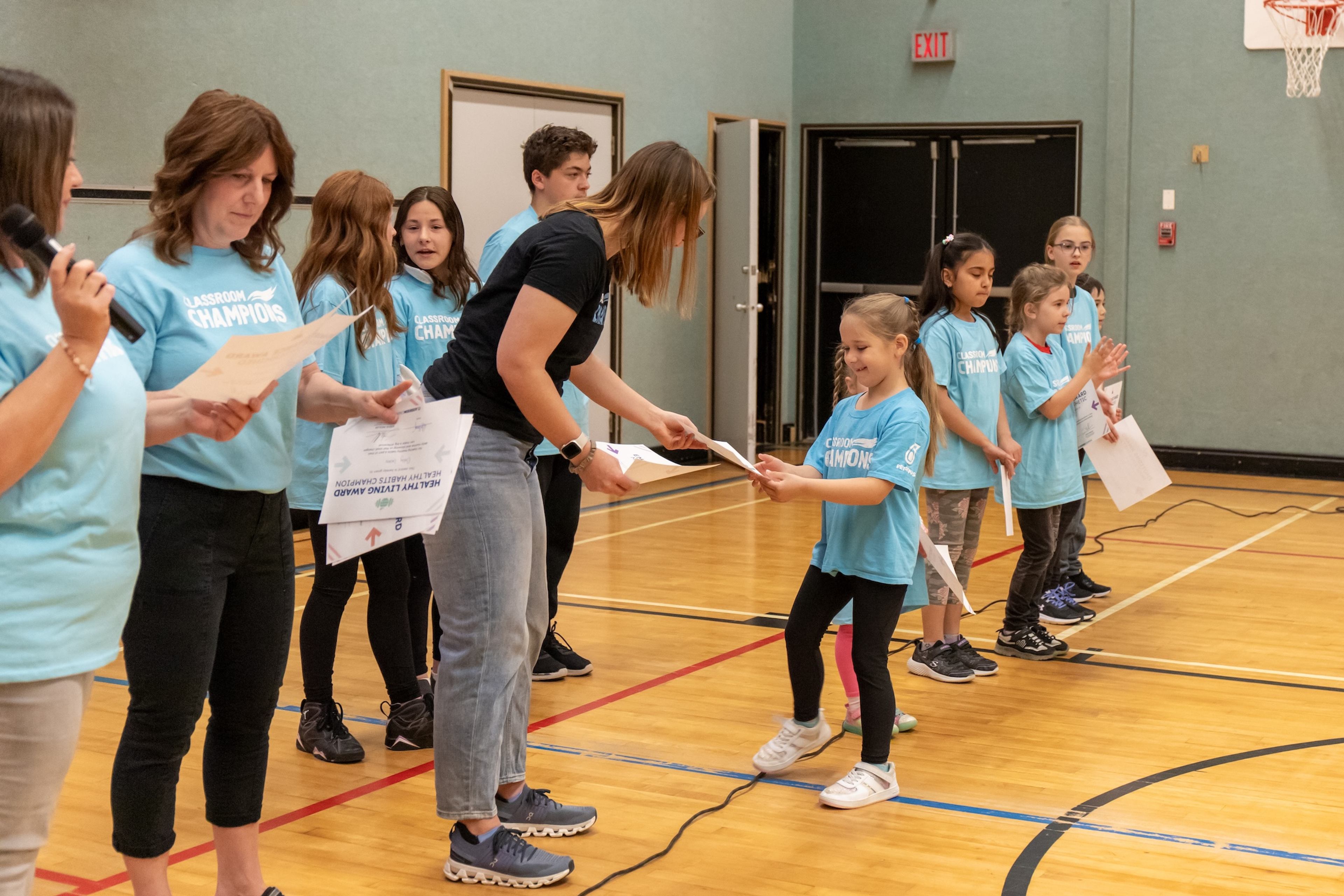 Students in Classroom Champions shirts receive award certificates during a school gym ceremony.