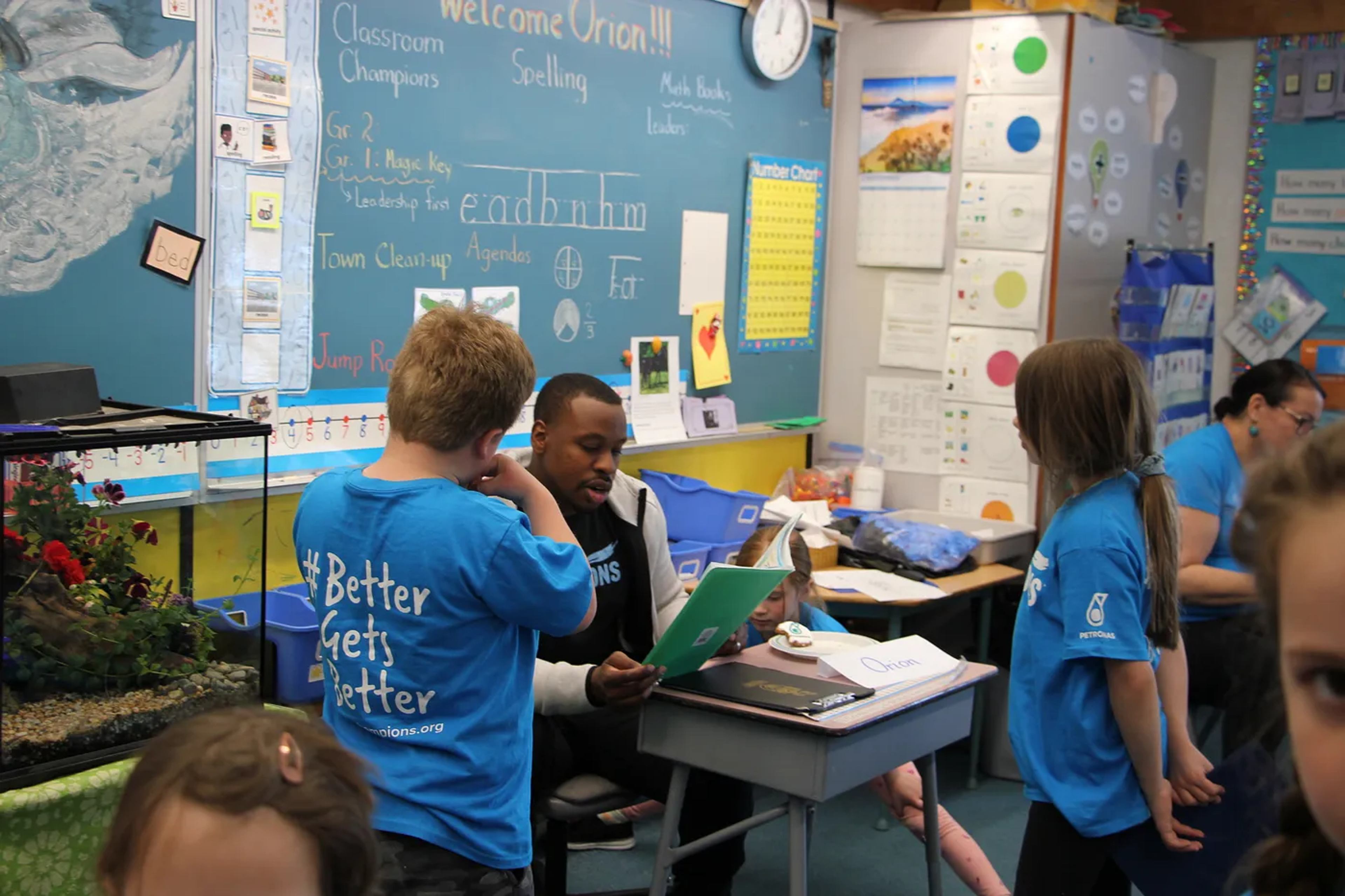 An athlete mentor sits with students wearing Classroom Champions shirts in a classroom decorated with a welcome sign for Orion.