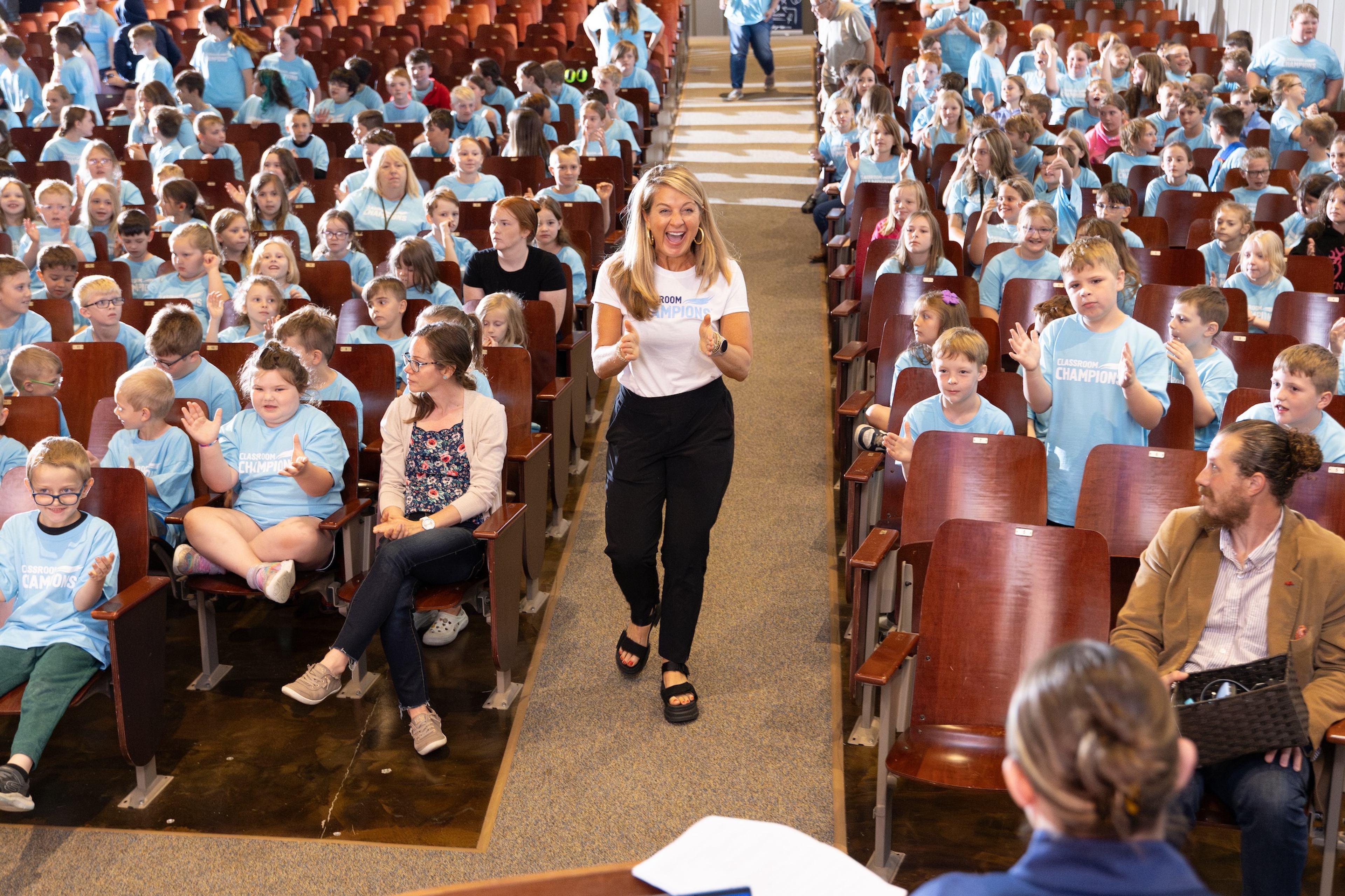 A Classroom Champions staff member walks an auditorium aisle giving thumbs up to cheering students in matching t-shirts.