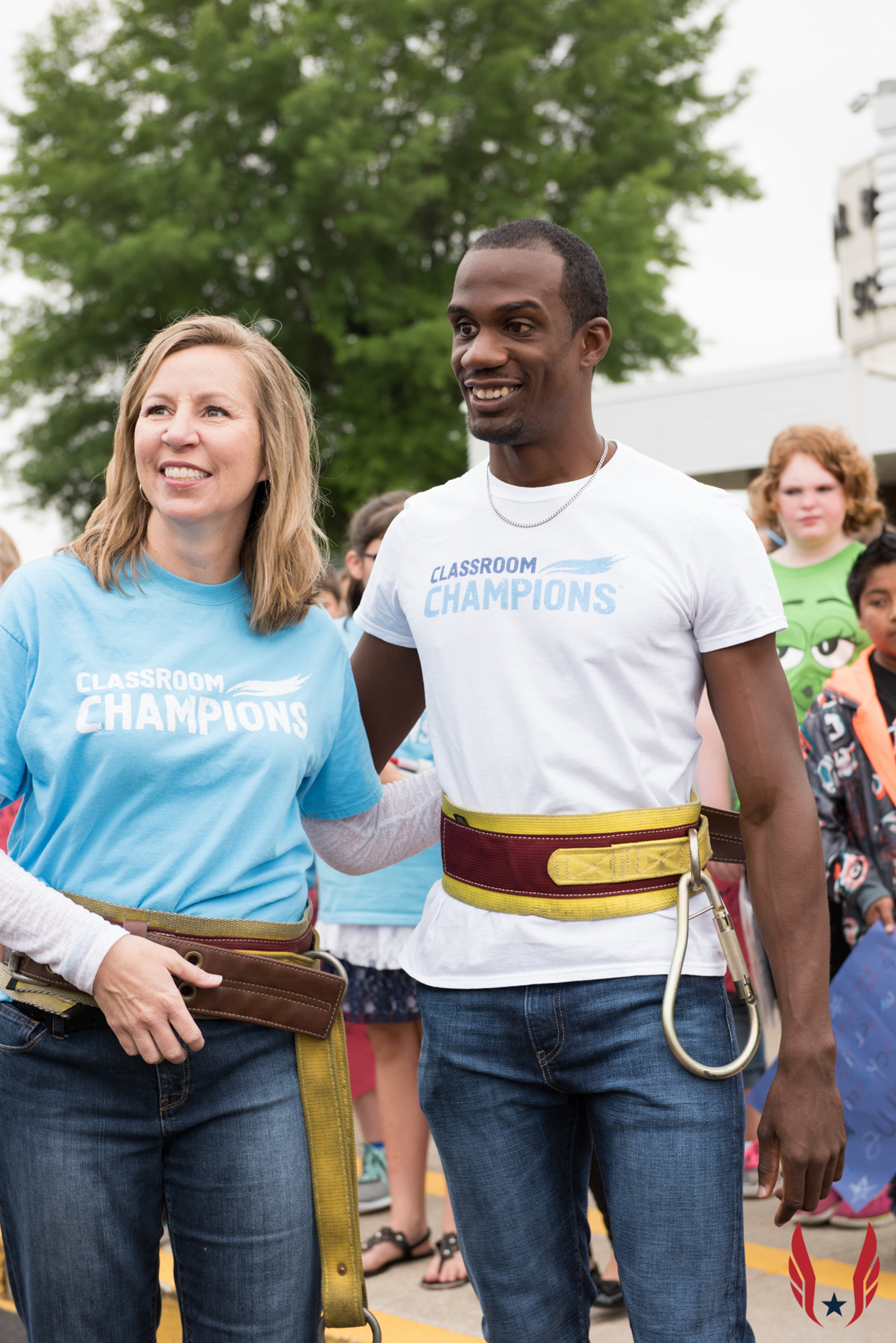 Two people wearing Classroom Champions t-shirts and safety harnesses stand together at an outdoor event.