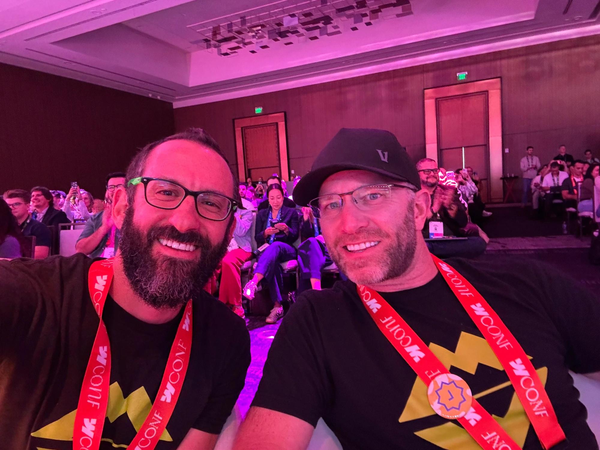 David (left) and Jesse (right) smiling for a selfie at Webflow Conf 2026, wearing Scaler branded shirts and conference lanyards with attendees seated behind them.
