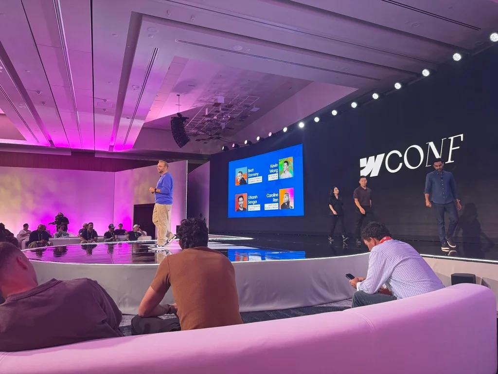 Presenters on stage at the Webflow Conf 2026 Builder Keynote, with the WCONF backdrop and the speaker lineup on screen, and attendees watching from lounge seating.