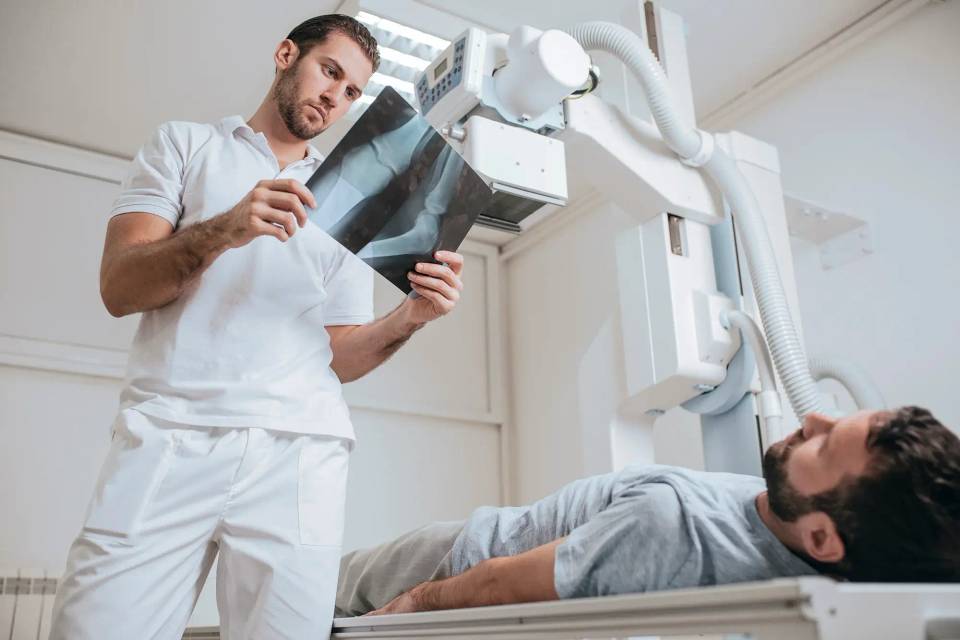 Radiology technician reviewing an X-ray image while a patient lies on an X-ray table.