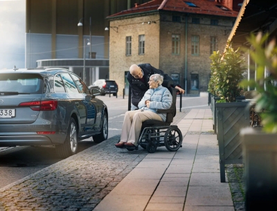 Gray car with Carony wheelchair transfer system, person in wheelchair beside caregiver preparing transfer into vehicle