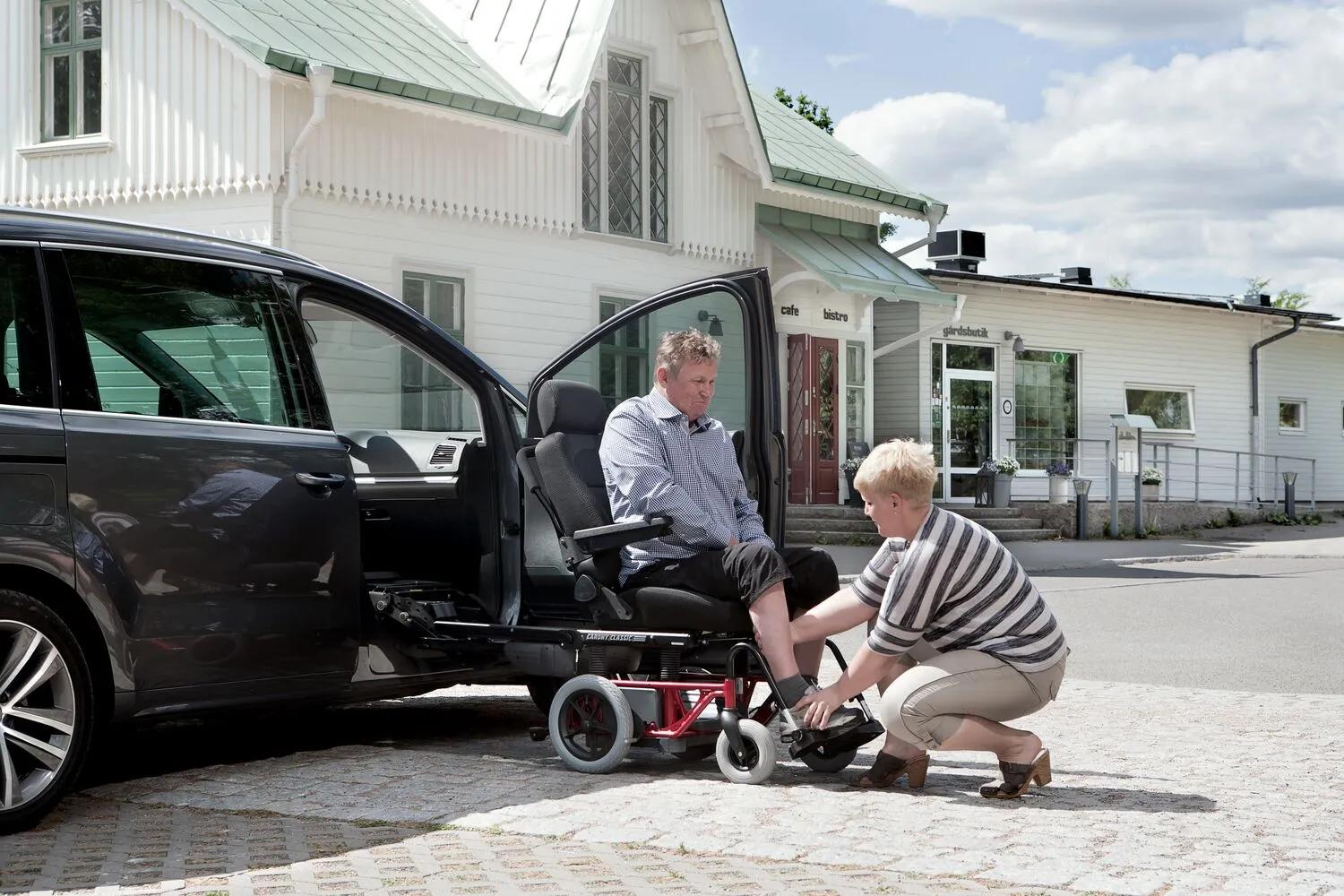 Gray car with Carony wheelchair transfer system, person in wheelchair assisted by caregiver adjusting footres