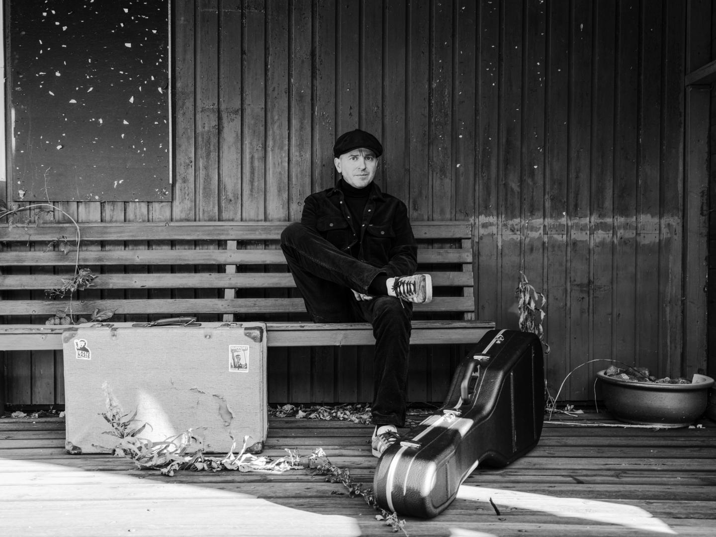 Bilde av Johnny Hide på ein benk, med gitarkasse foran seg.
