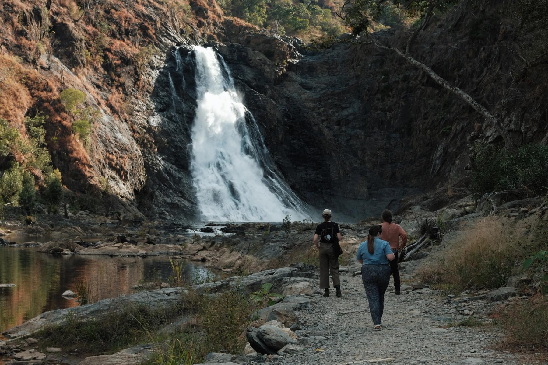 Three people walk towards a large cascading waterfall over rocky cliffs into a calm pool