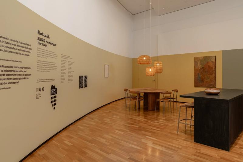 Curved wall with text about the After the Rain exhibition, spaces featured dining table and chairs, woven pendant lights and a kitchen bench