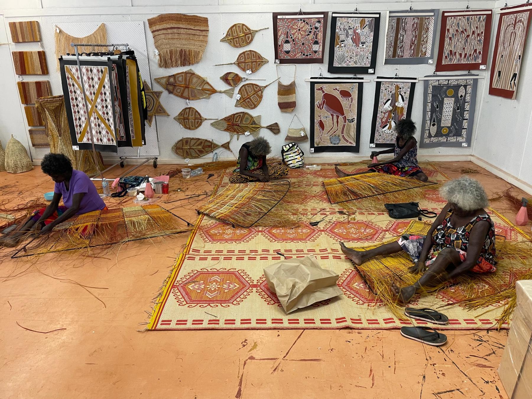 Four women sitting on the floor weaving pendants, artwork is on the surrounding walls.