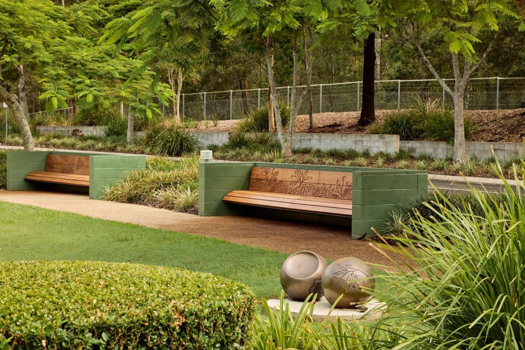 Garden with grass, trees and hedges, with detailed timber benches and a gumnut sculpture in the foreground