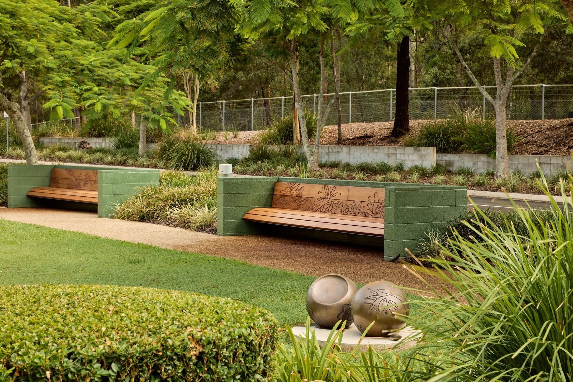 Garden with grass, trees and hedges, with detailed timber benches and a gumnut sculpture in the foreground